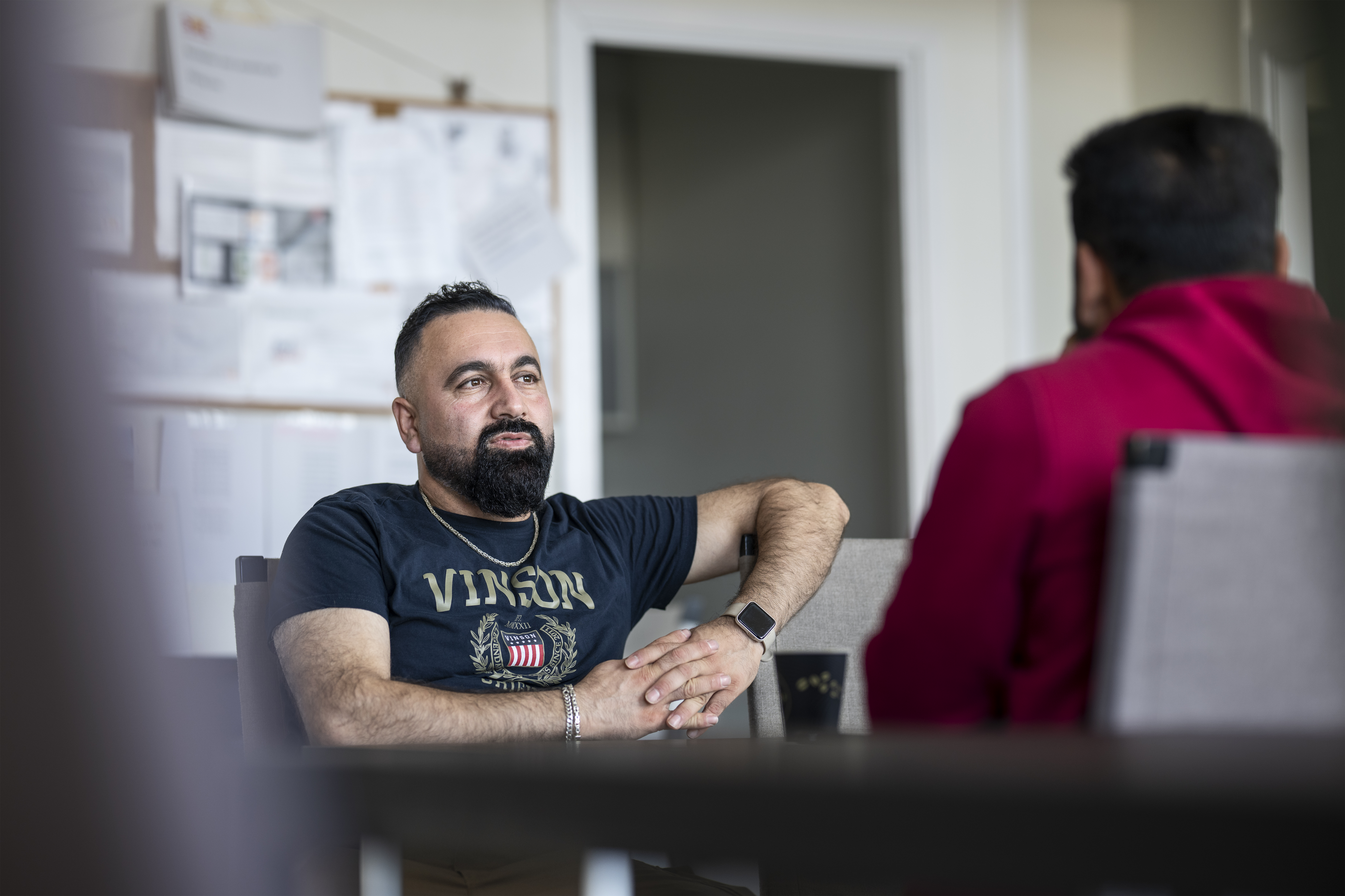 Två män sitter och pratar i ett lunchrum med papper uppsatta på en anslagstavla i bakgrunden.