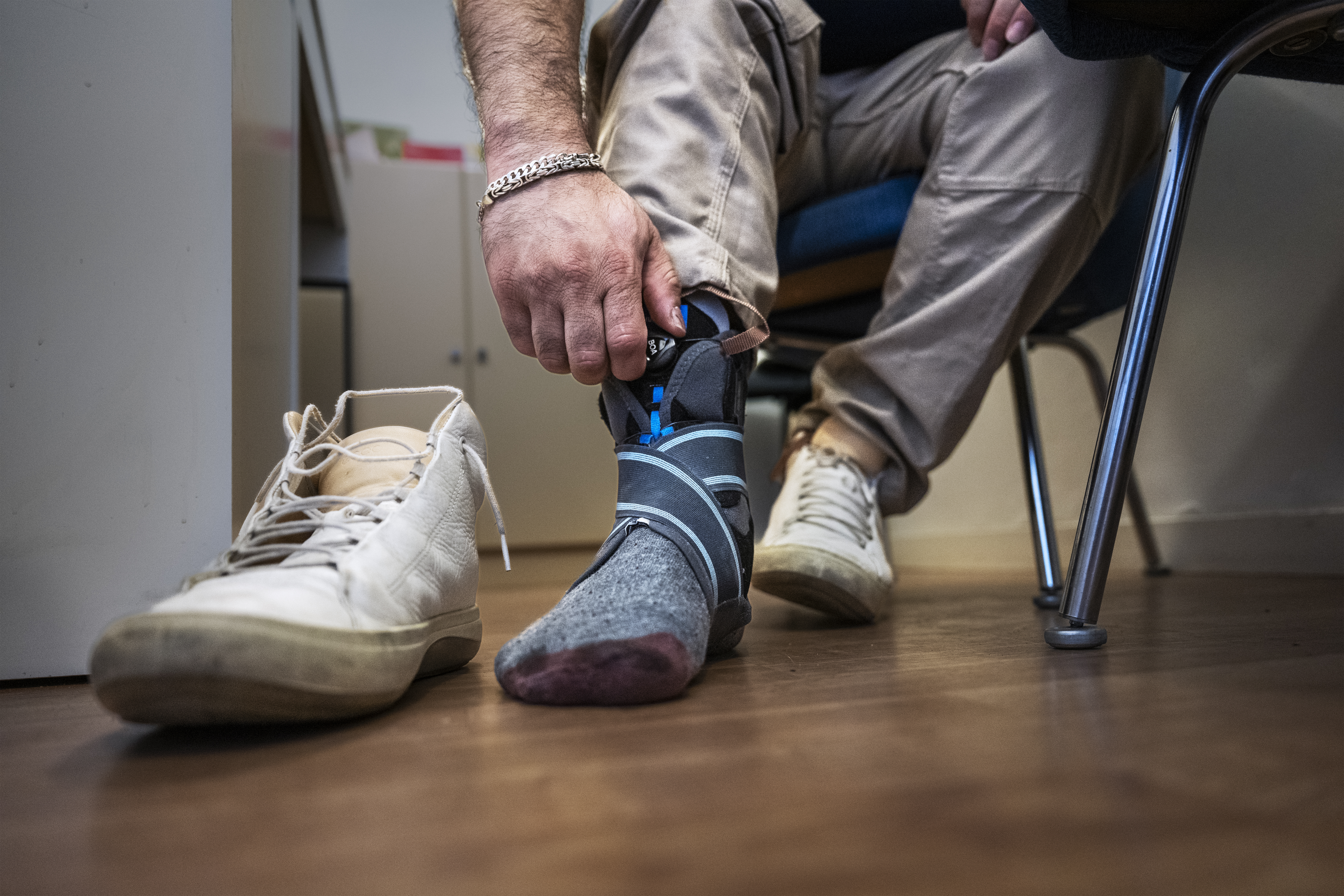 En person justerar ett stödbandage över sin strumpklädda fot. En gymnastiksko står bredvid på golvet.