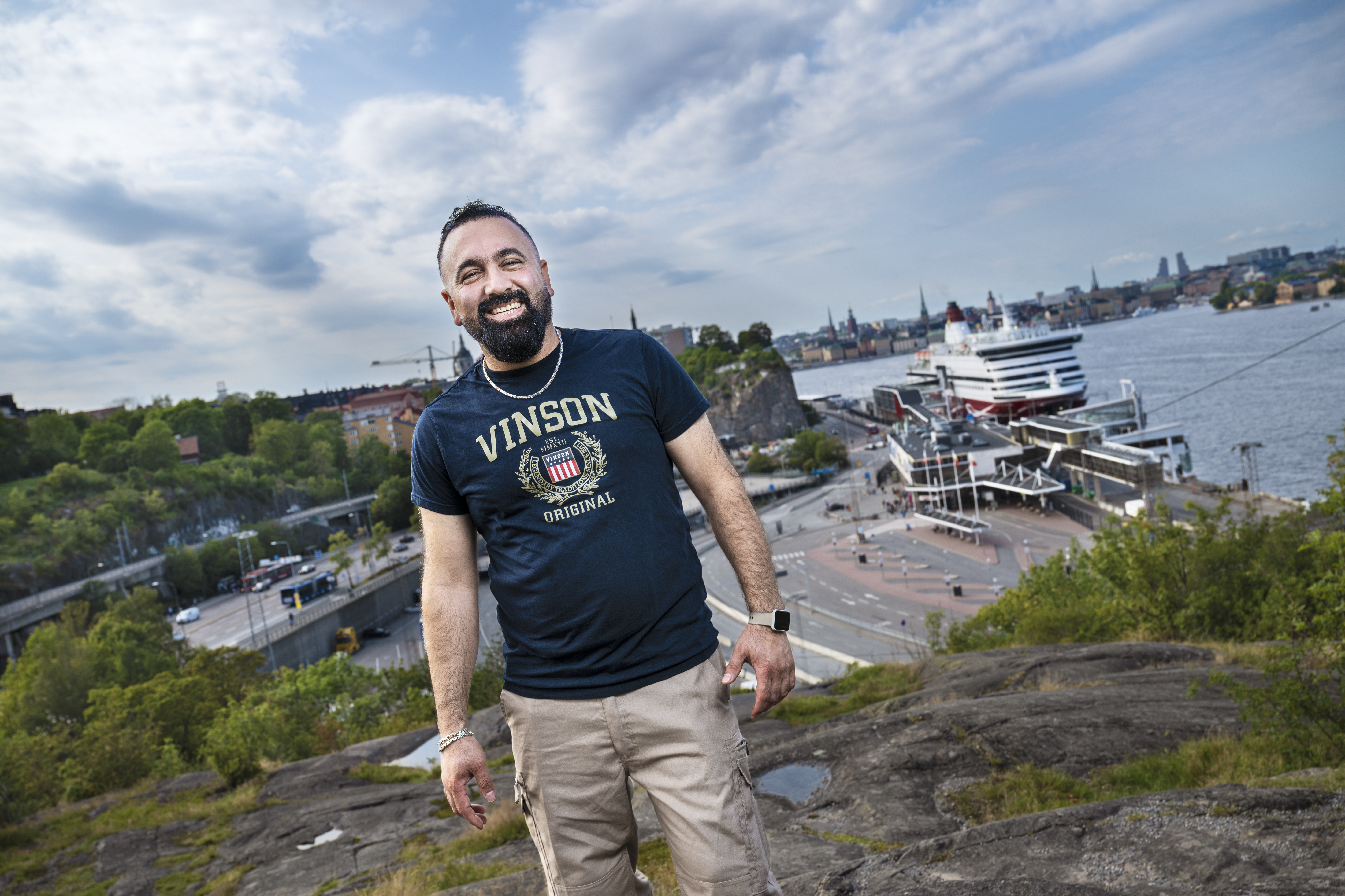 En skäggig man står på en klippa. Bakom honom syns en hamn med ett kryssningsfartyg och en stadsbild i bakgrunden.