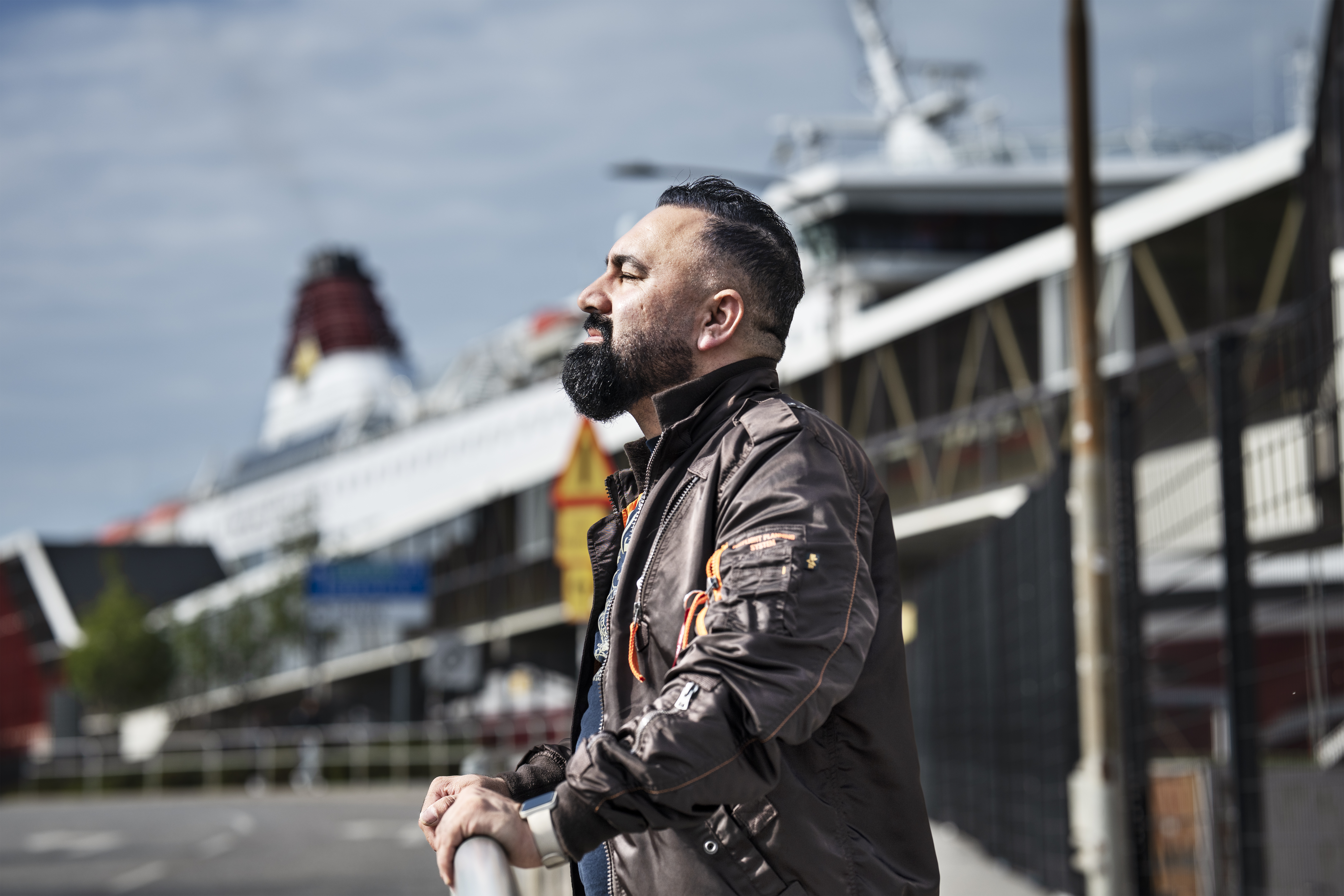 En man med skägg och mörkt hår står utomhus och lutar sig mot ett räcke med slutna ögon. Ett kryssningsfartyg syns bakom.