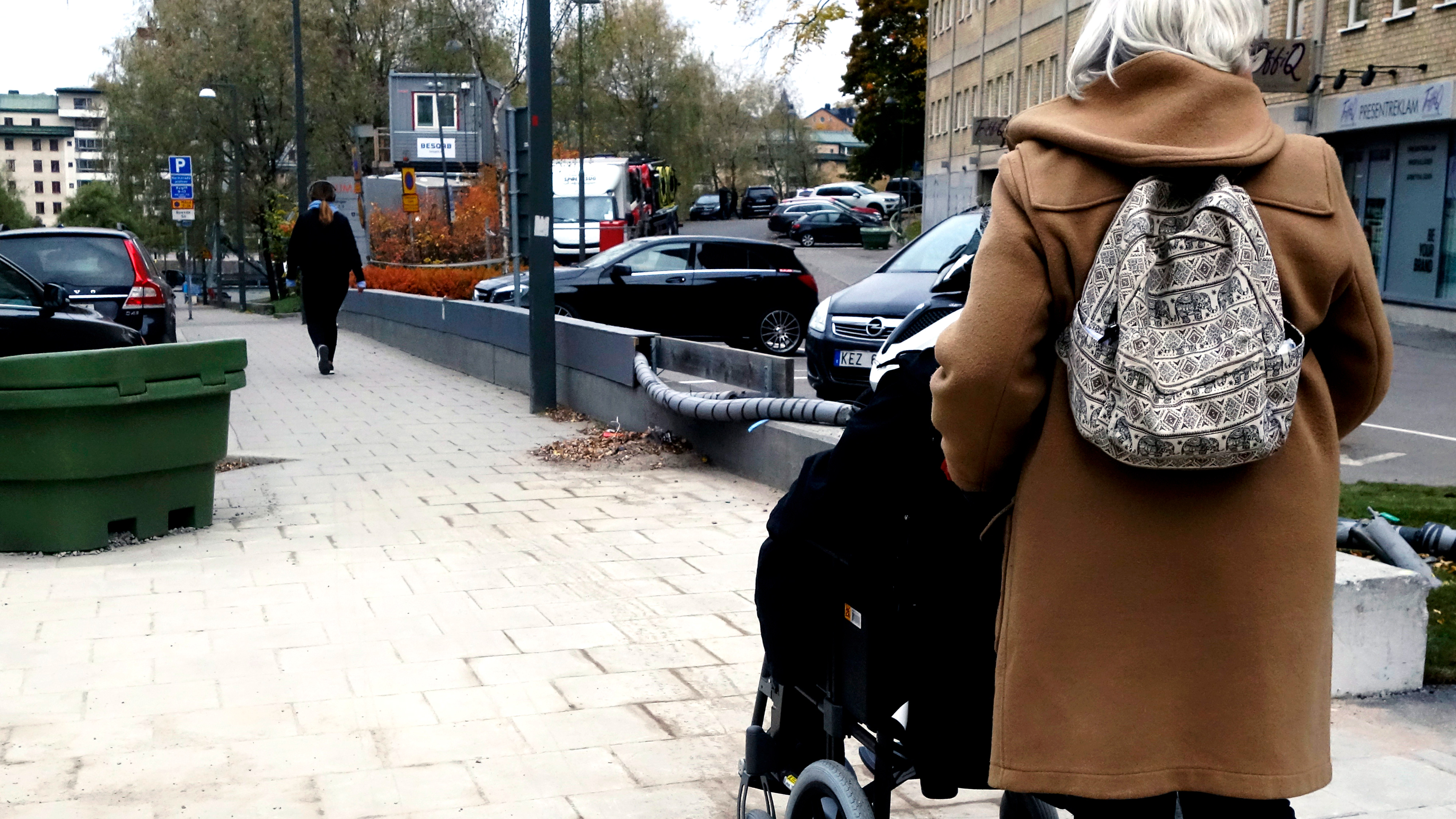 En äldre vuxen med beige rock och mönstrad ryggsäck skjuter en person i rullstol längs en trottoar i staden.