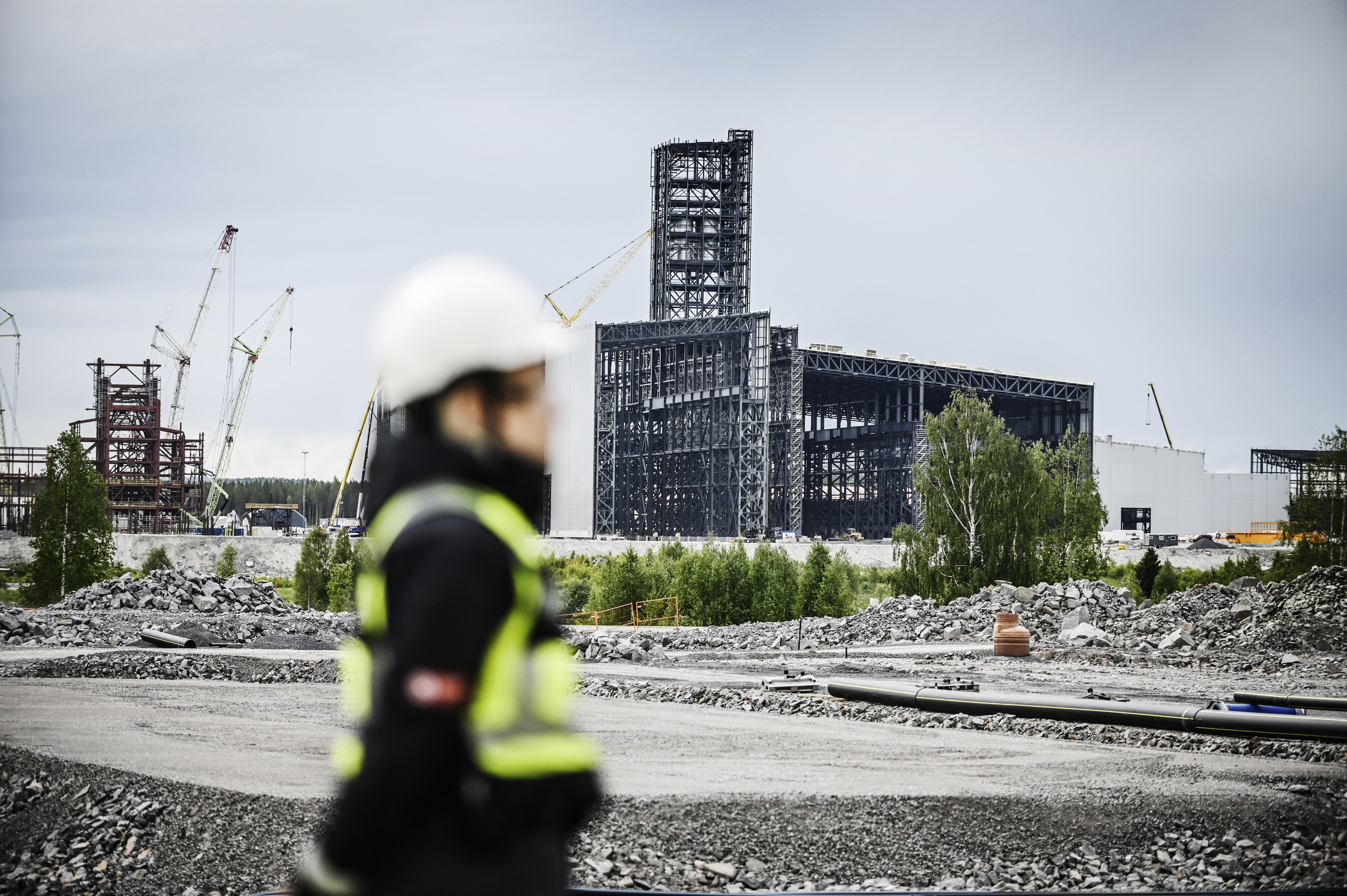 En byggarbetare med skyddsutrustning står i förgrunden, bakom syns en industribyggnad under uppförande och kranar.