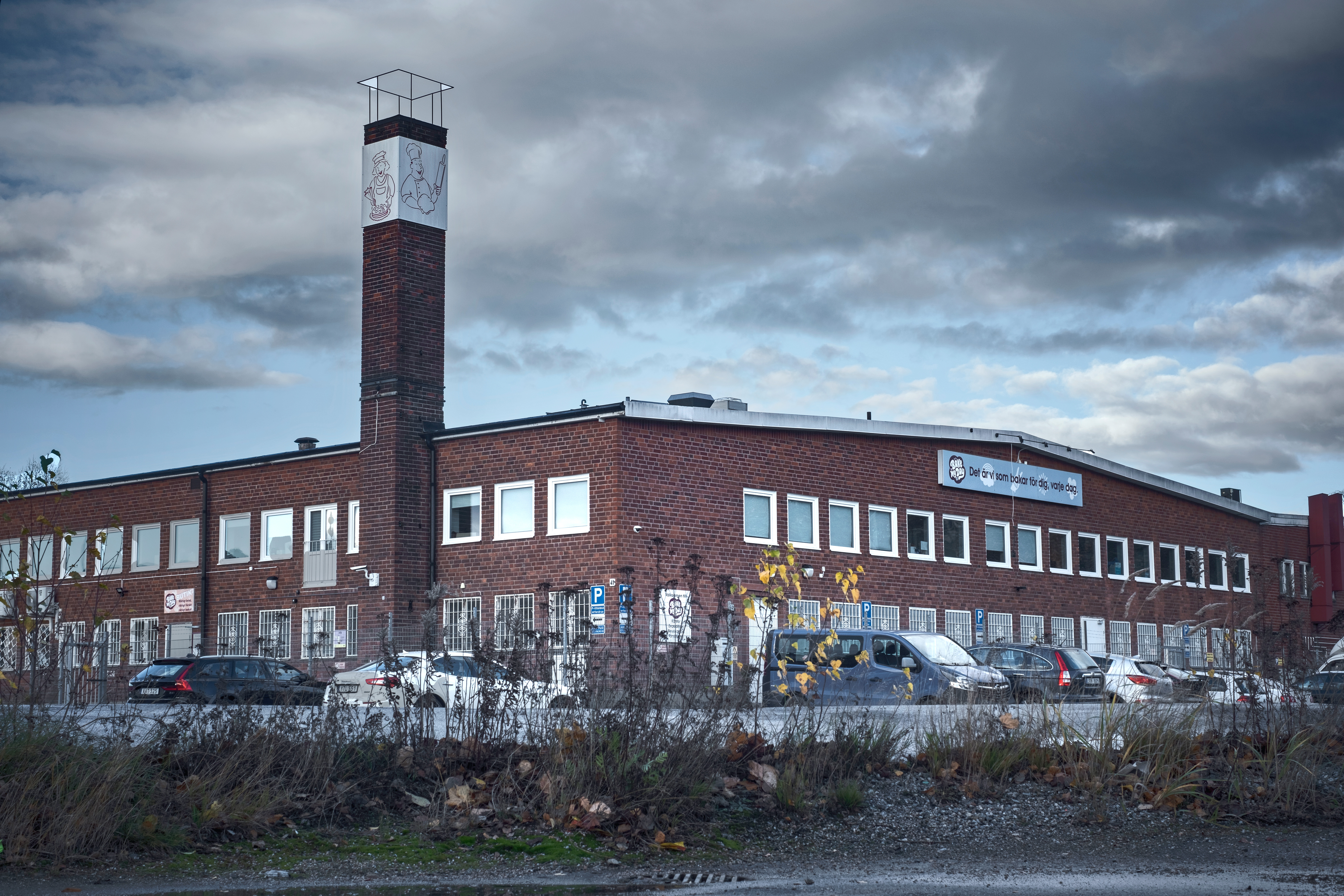En tegelbyggnad med två våningar och en hög skorsten, flera bilar står parkerade framför, himlen är molnig.