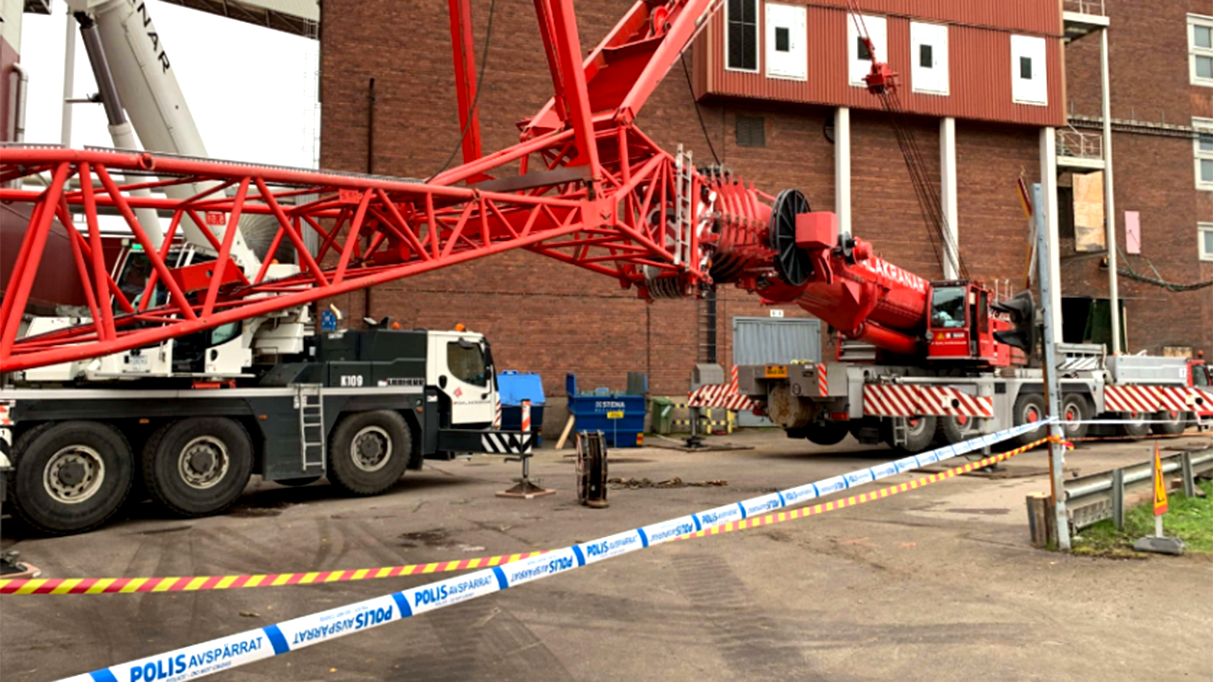 En röd kran har vält vid en byggarbetsplats bredvid en tegelbyggnad, avspärrningsband och utryckningsfordon syns.