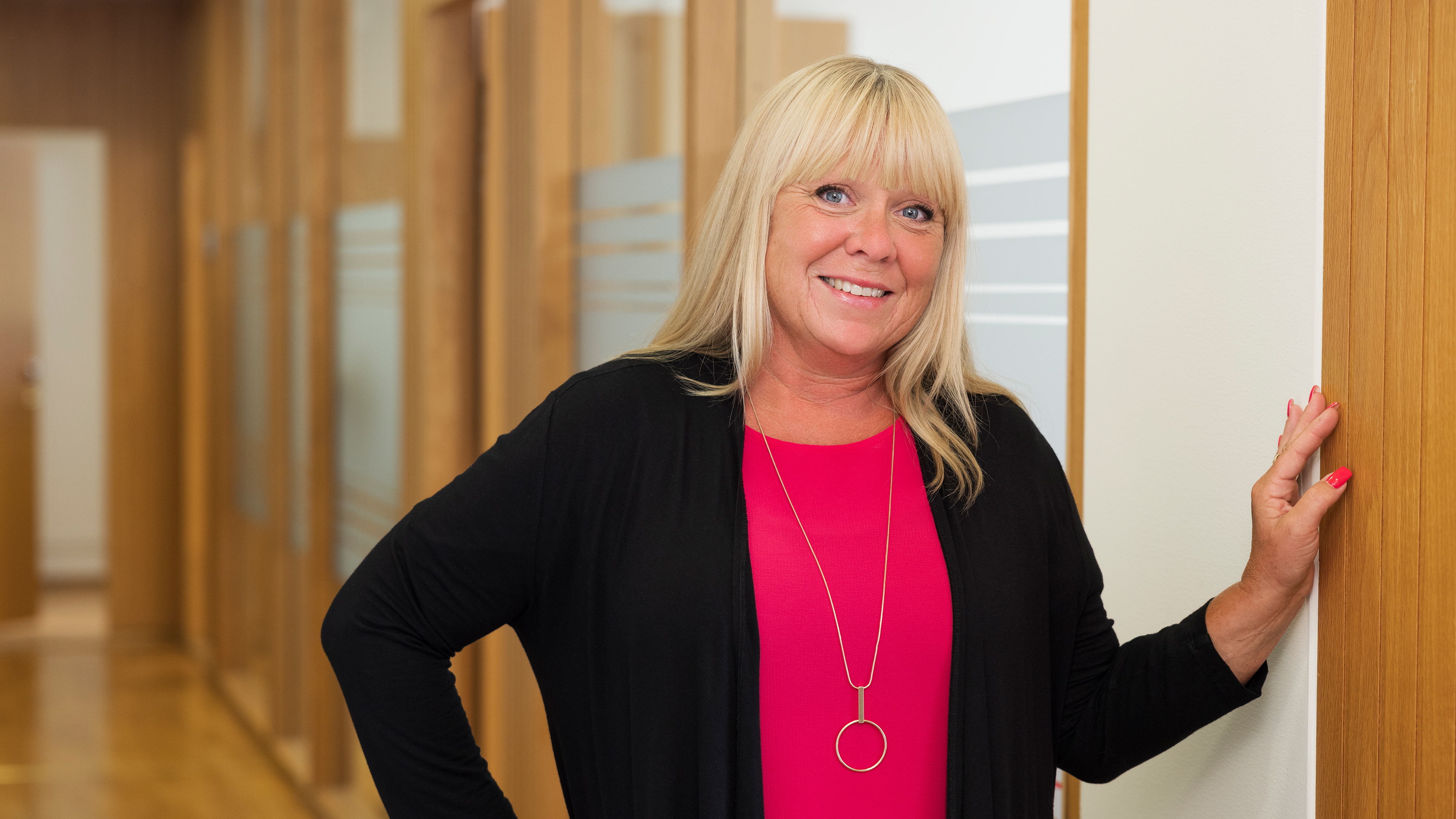 En kvinna med blont hår har på sig en rosa topp och svart kofta. Hon står inomhus vid en trävägg och ler mot kameran.