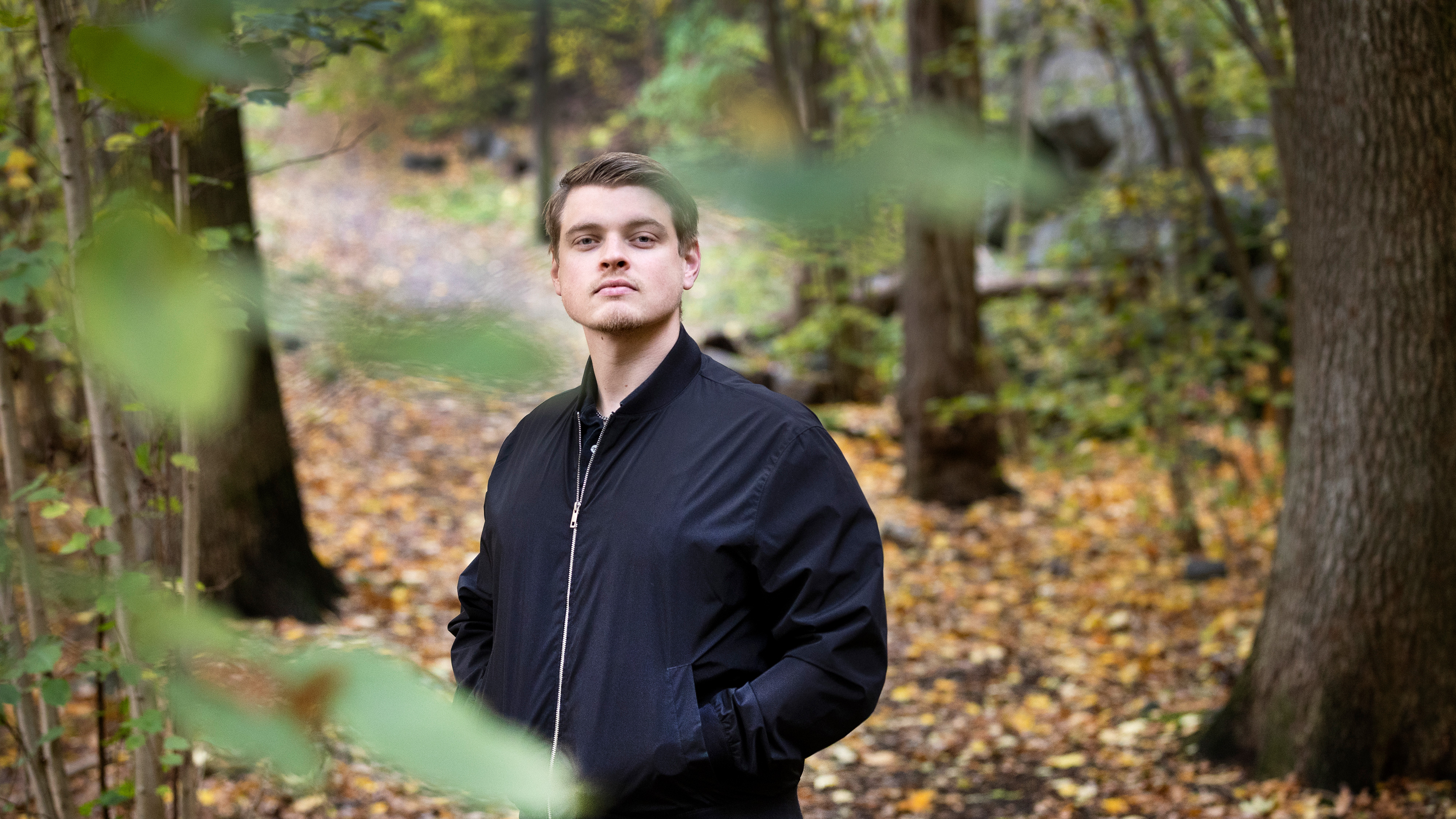 Tobias Eng Strömberg i svart jacka står i en skog med löv på marken och gröna växter framför.