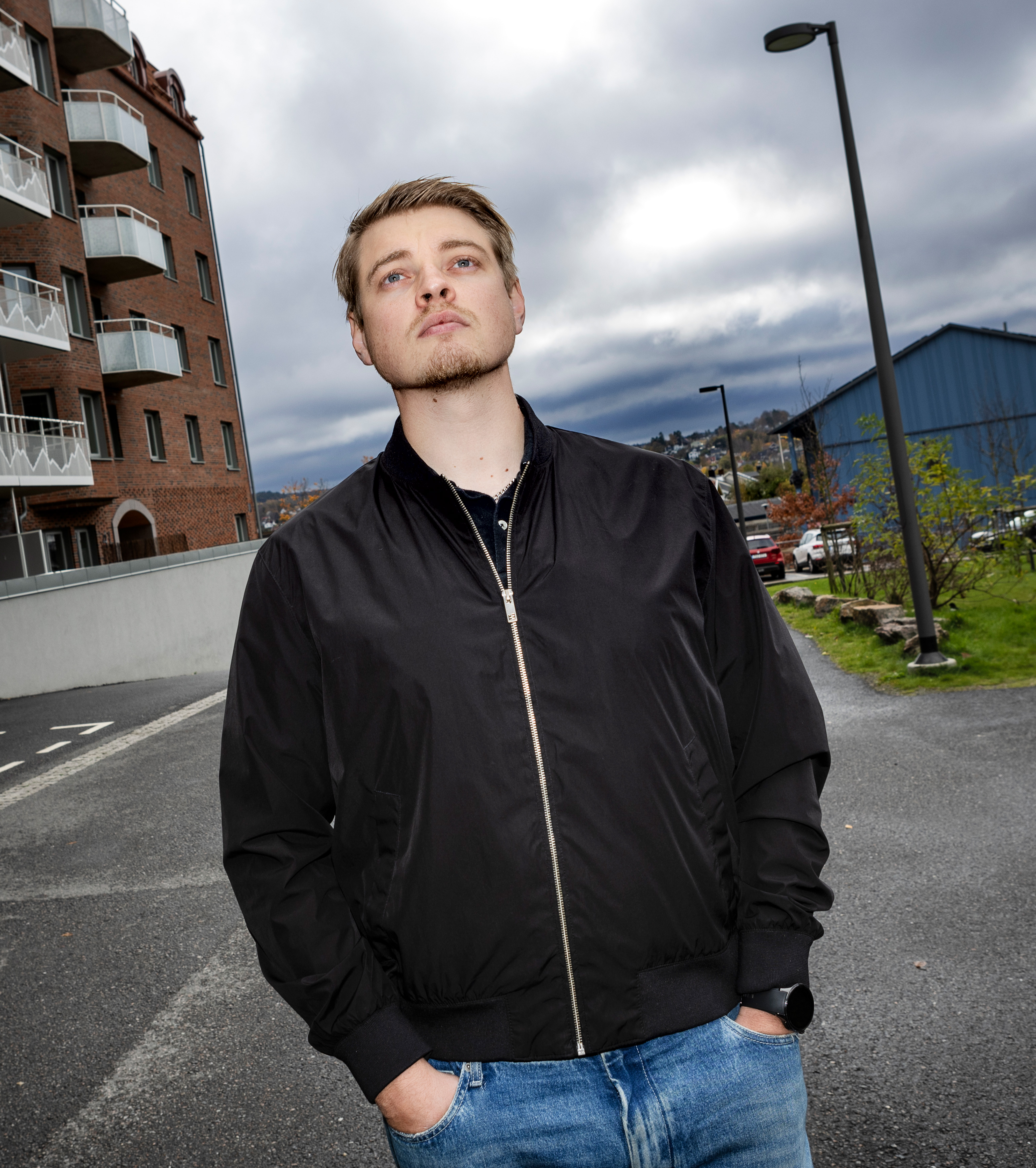 Tobias Eng Strömberg med svart jacka och blå jeans står utomhus med händerna i fickorna, nära ett hyreshus och en gatlykta under molnig himmel.