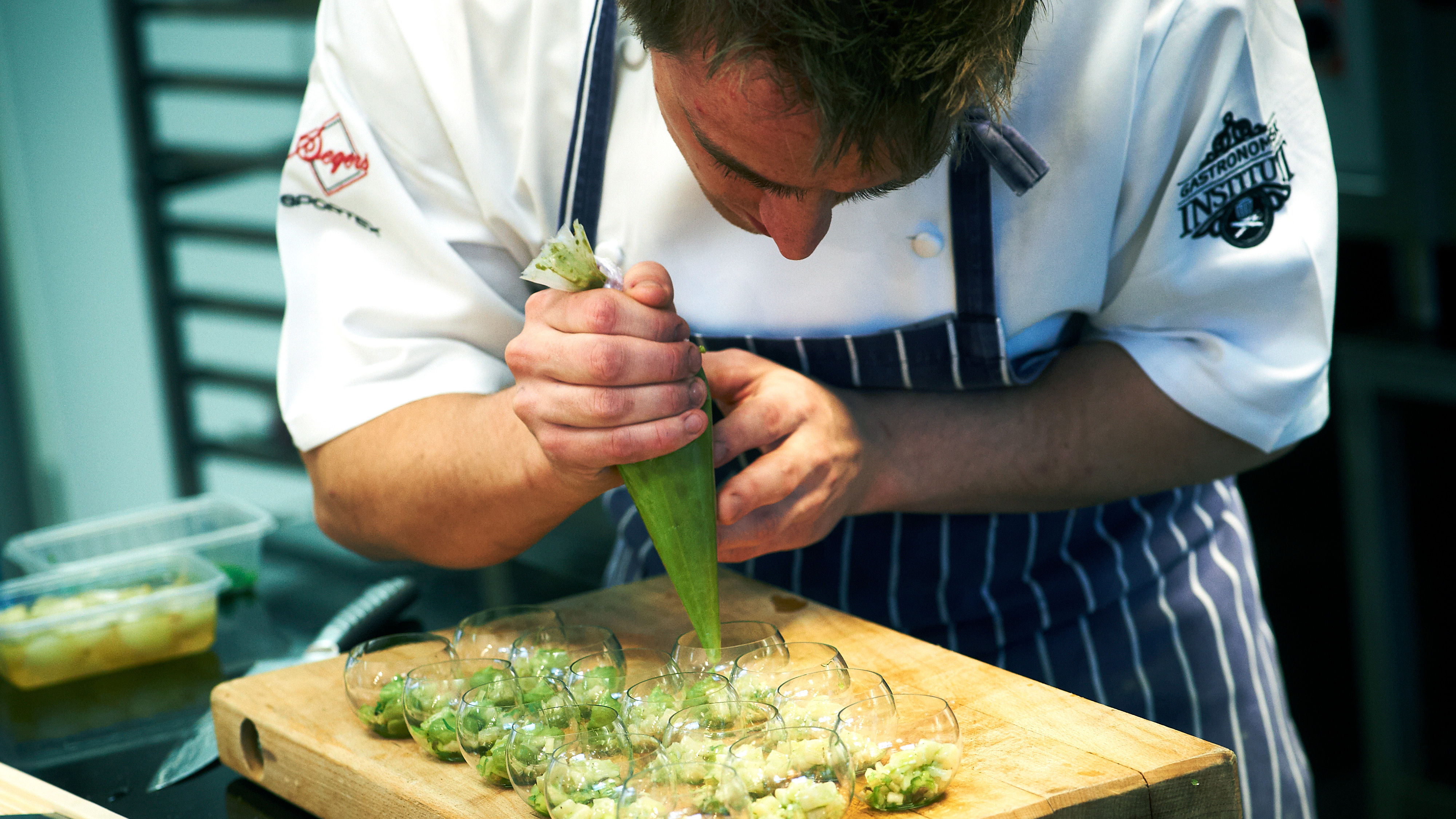 En kock i uniform spritsar en grön blandning i små glasbägare som står på en träskärbräda.