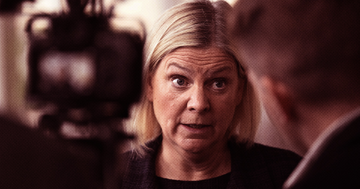 En kvinna med blont hår pratar med någon i ett svagt upplyst rum, kamerautrustning syns i förgrunden.