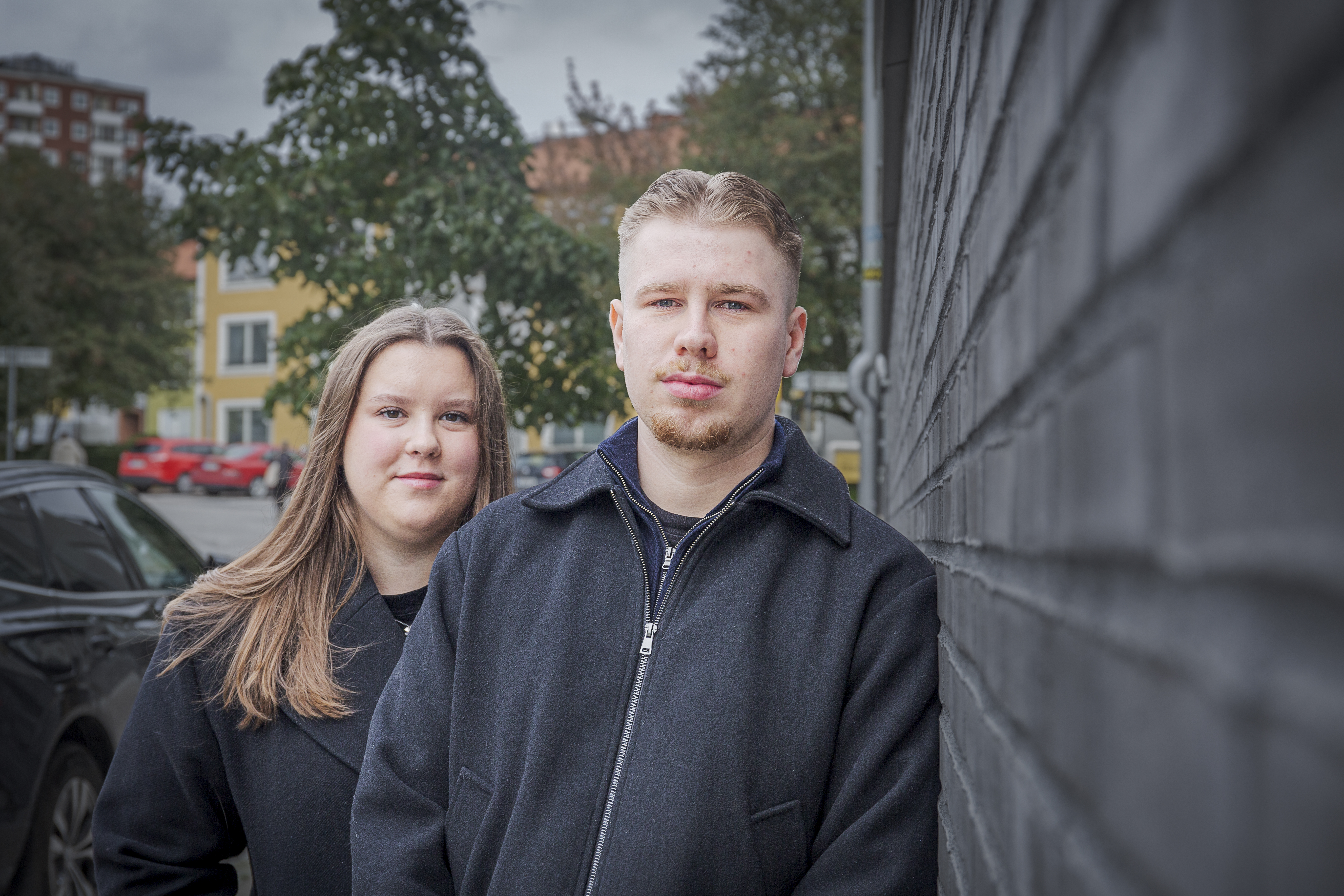 Flickvännen My och Rasmus Ström står utomhus i mörka rockar. Mannen står längst fram mot en tegelvägg, kvinnan står lite bakom. Hus och träd syns i bakgrunden.