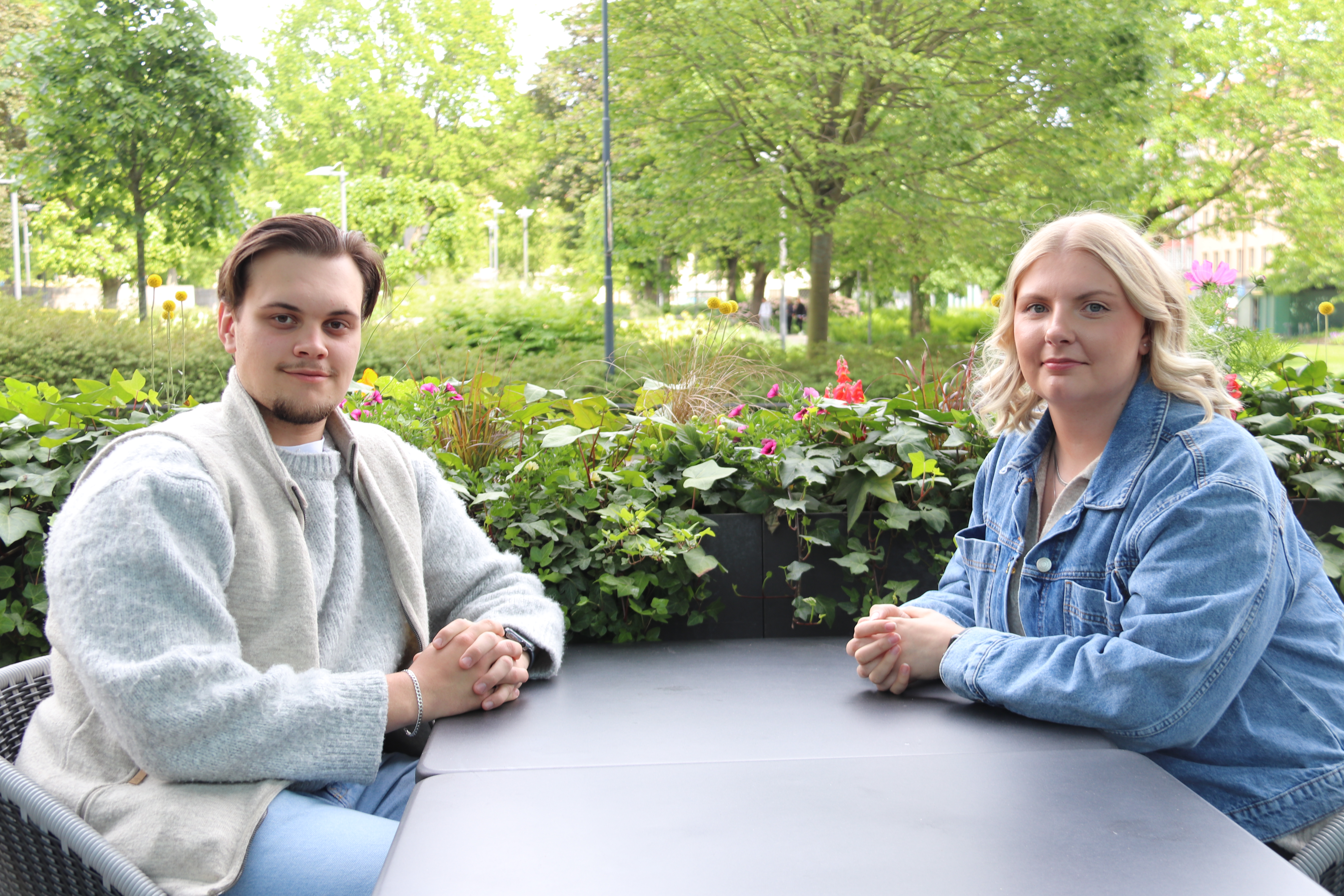 Två personer sitter mittemot varandra vid ett bord utomhus, vända mot kameran med händerna på bordet.