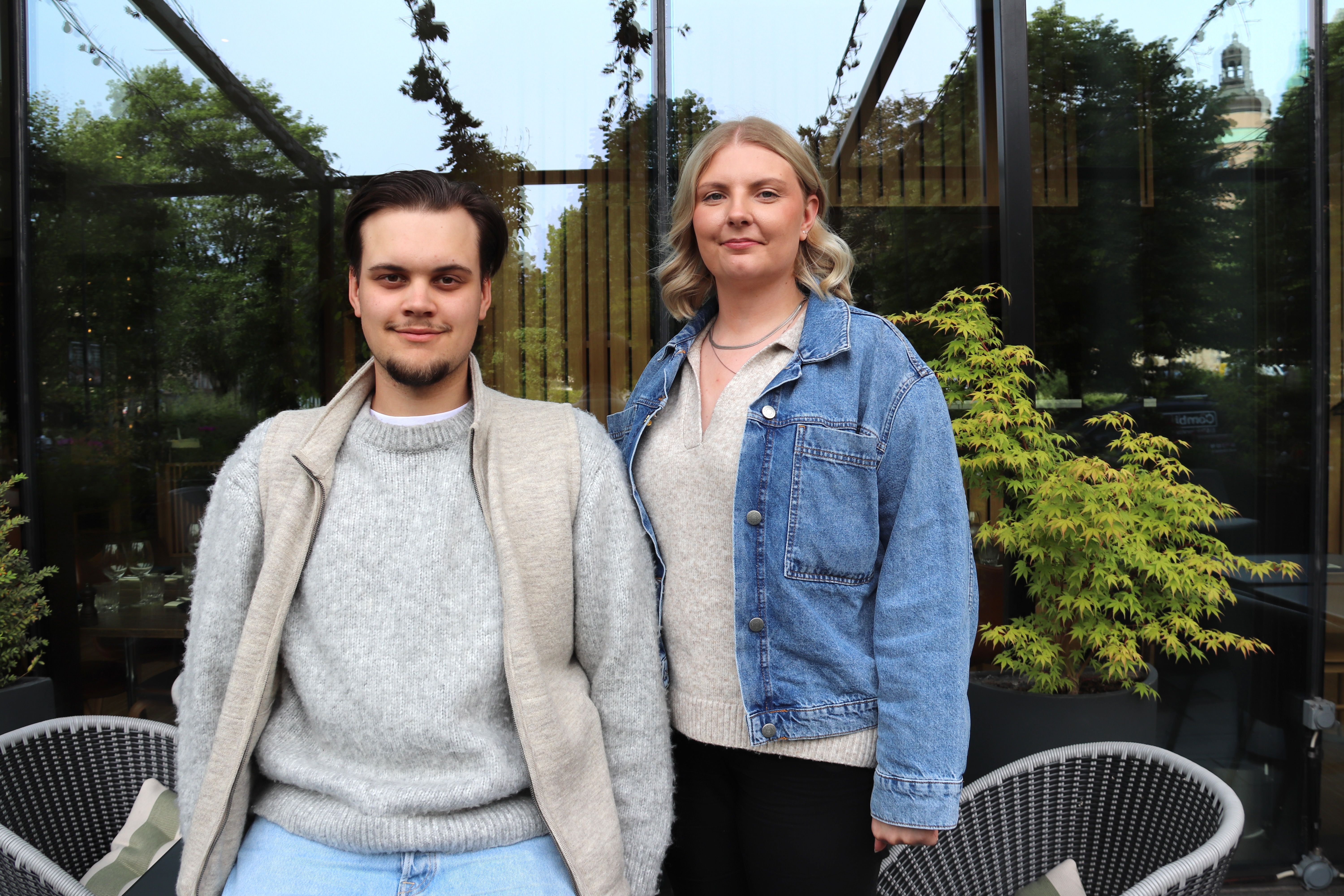 En man och en kvinna i tröjor och jackor står tillsammans utomhus mot bakgrund av växter och glasfönster.