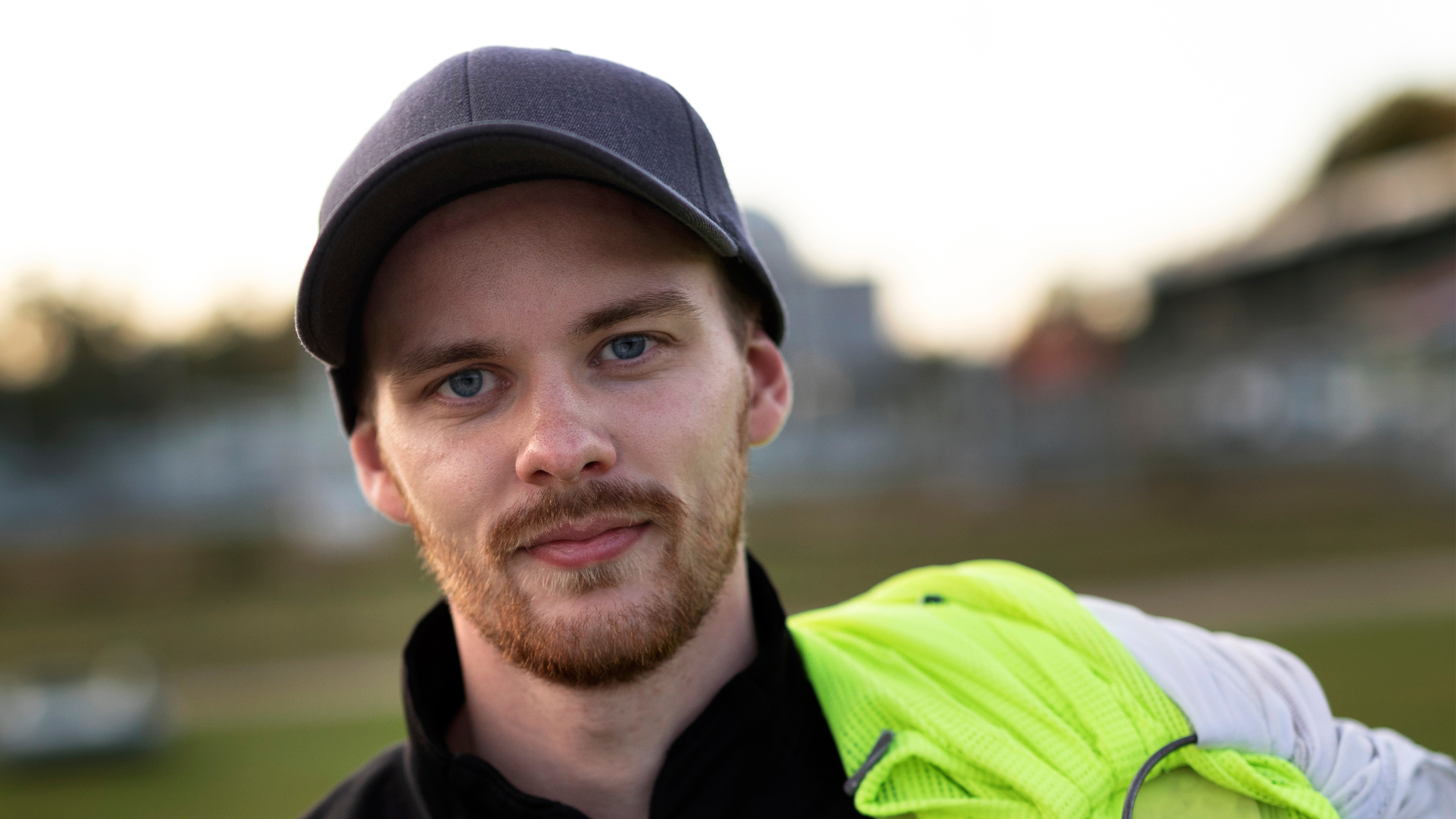 En man med kort skägg och blå ögon har en mörk keps och håller en neongul jacka över axeln, står utomhus.