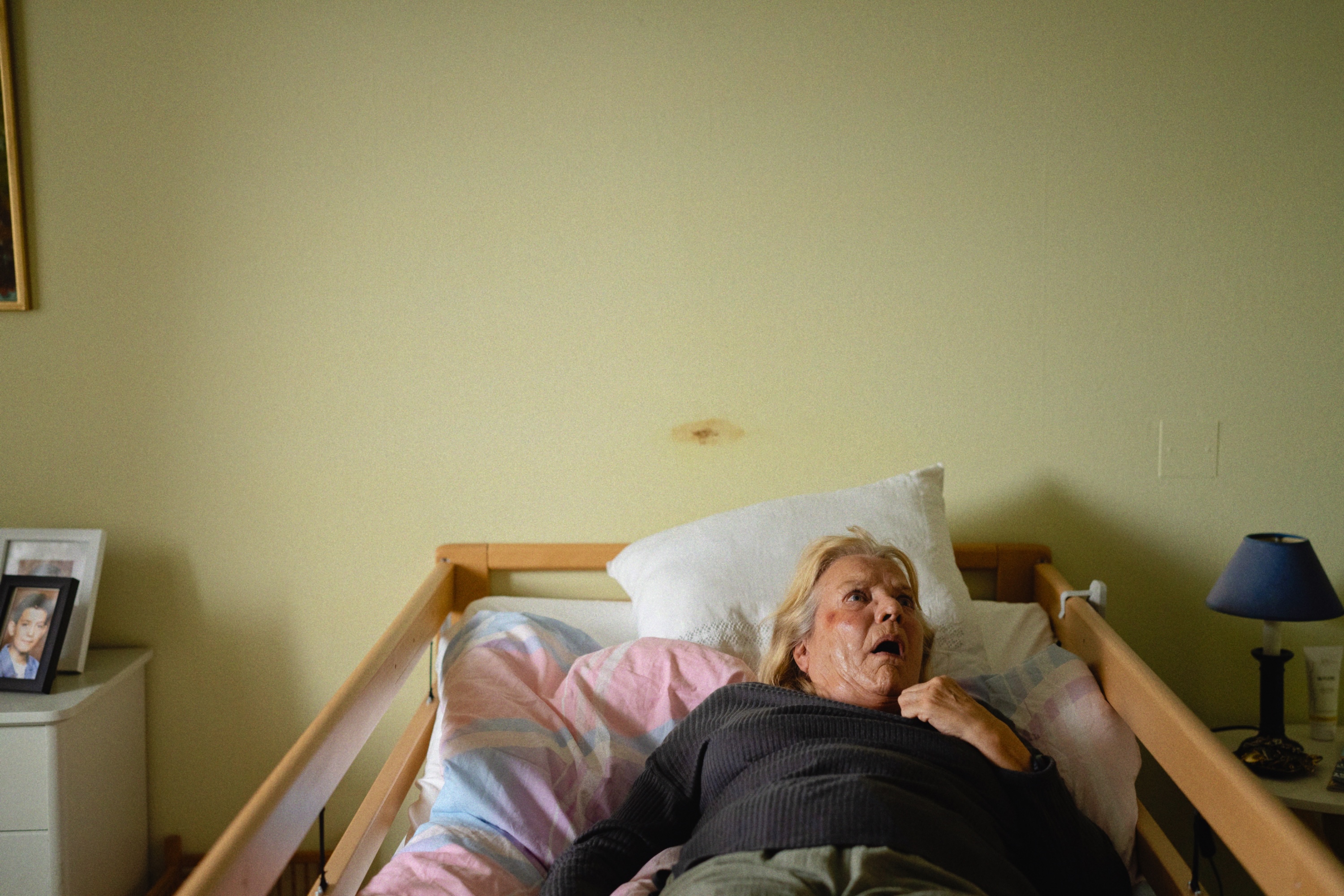 En äldre kvinna ligger i en träsäng på ett äldreboende, ser rädd ut med ena handen på bröstet. Ett inramat foto, lampa och byrå syns bredvid sängen.