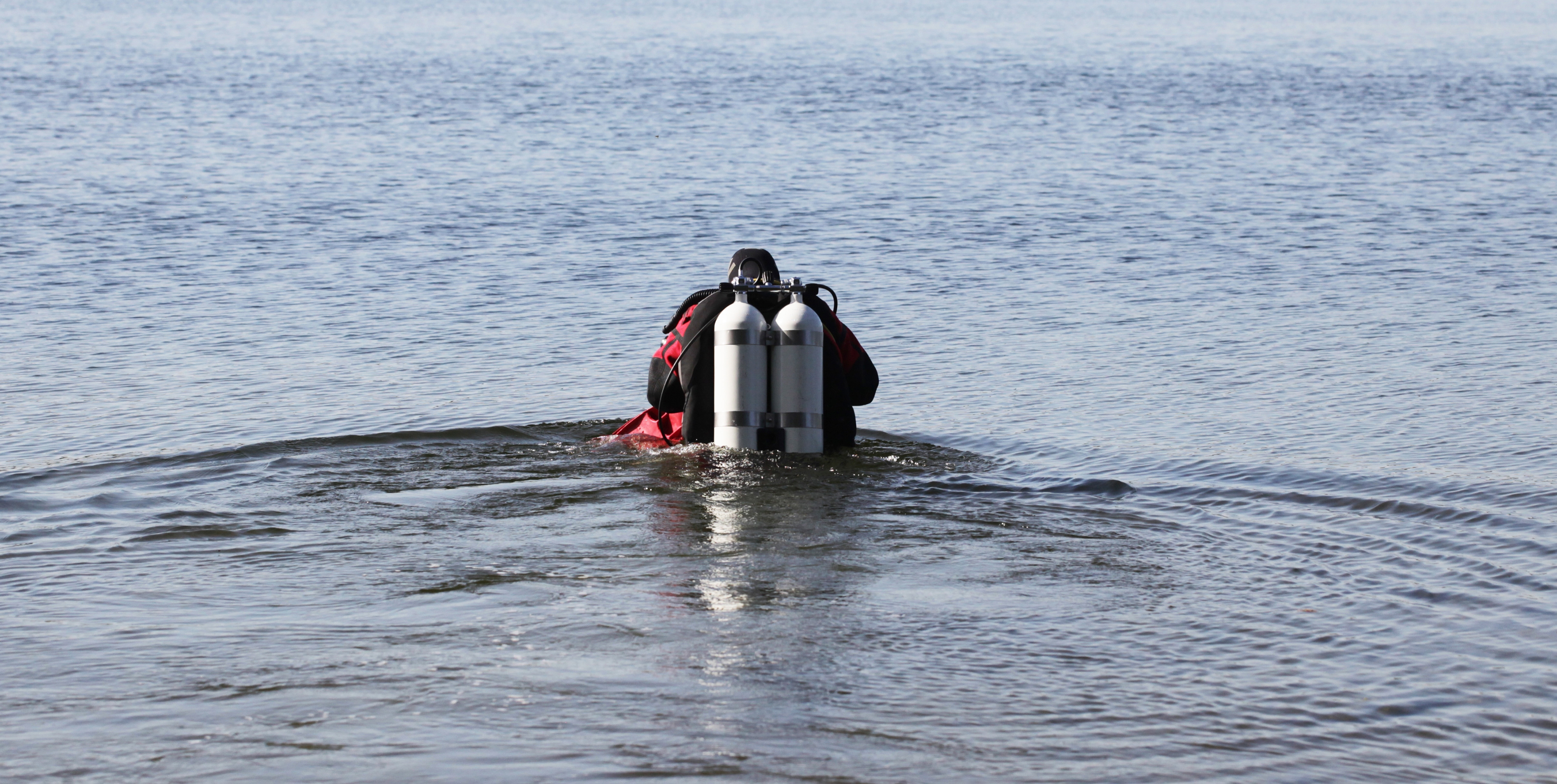 En dykare i våtdräkt och med två lufttuber står i grunt vatten, vänd bortåt och förbereder sig för att dyka i vattnet.