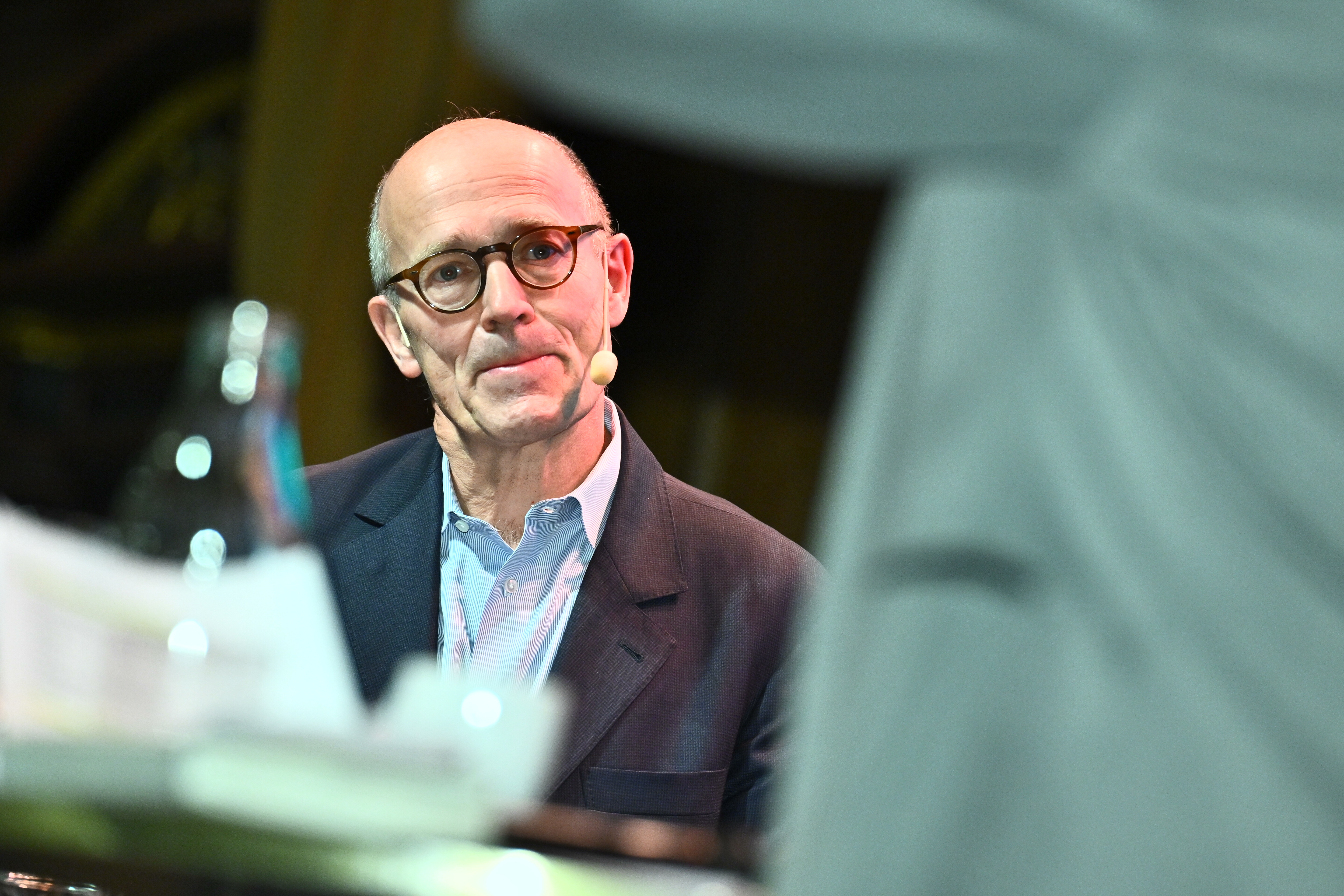 Harald Mix med glasögon och kavaj på en scen med ett mikrofonheadset. En flaska och papper syns i förgrunden.