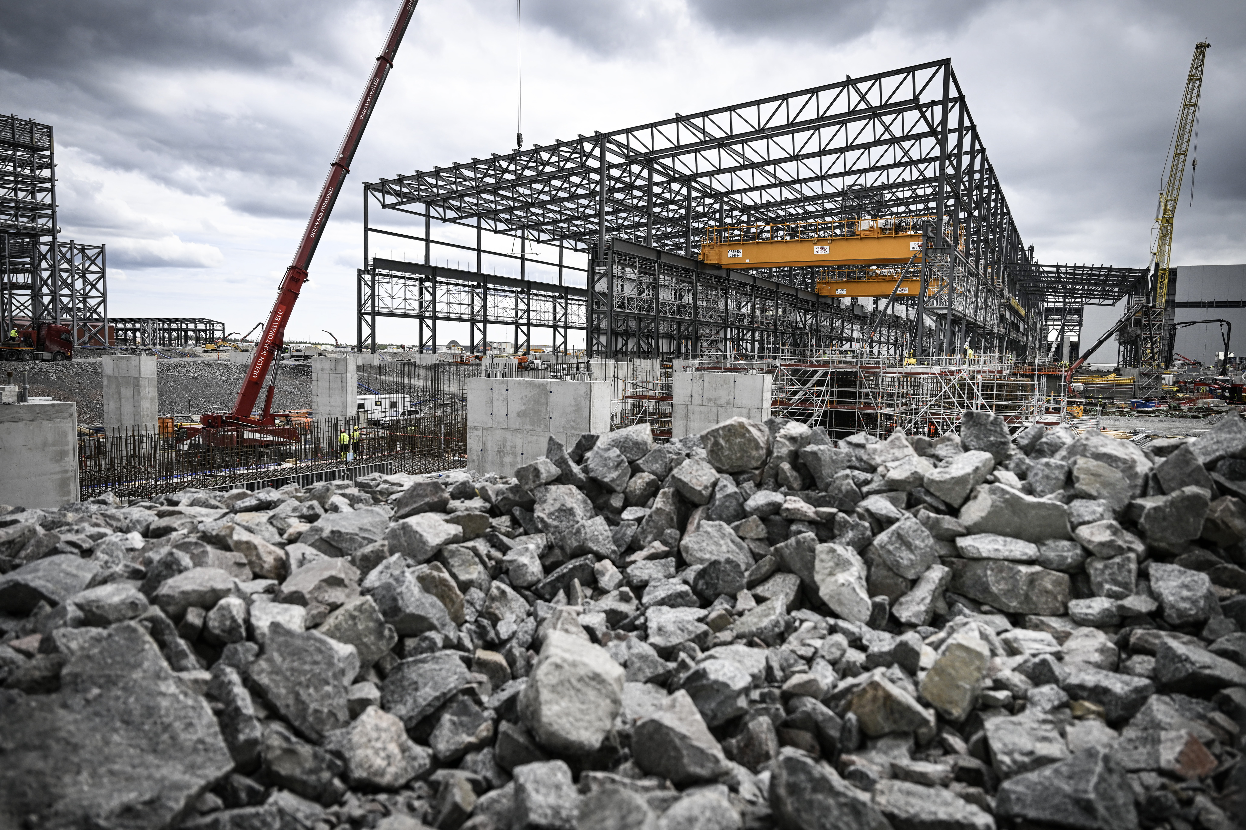 En byggarbetsplats med stålkonstruktioner, kranar och betongpelare samt en hög med sten i förgrunden under molnig himmel.