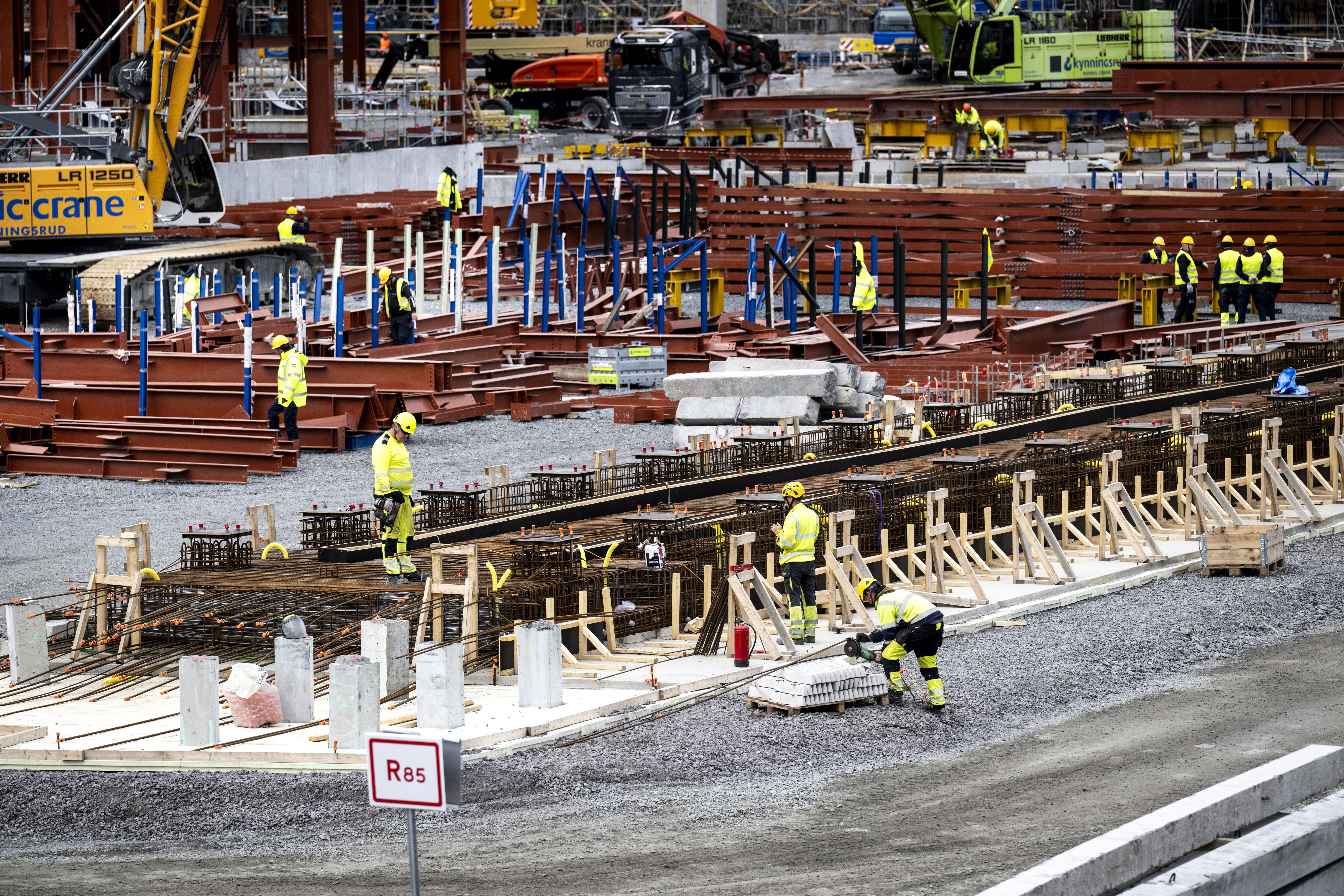 Byggnadsarbetare med skyddsutrustning monterar stålkonstruktioner och material på en byggarbetsplats med maskiner i bakgrunden.