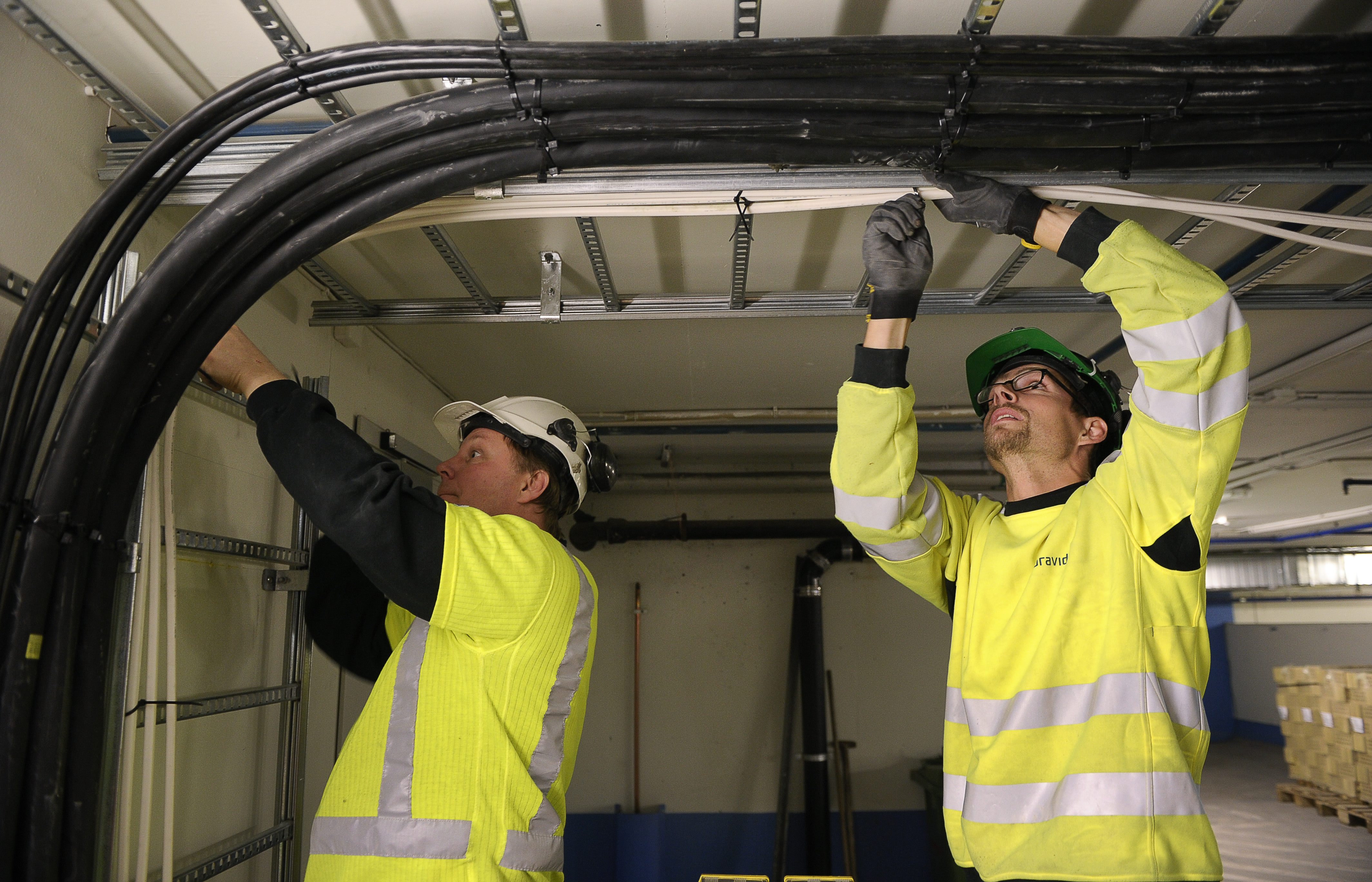 Två arbetare med varselkläder och hjälmar installerar eller justerar svarta kablar på en kabelställning i taket.