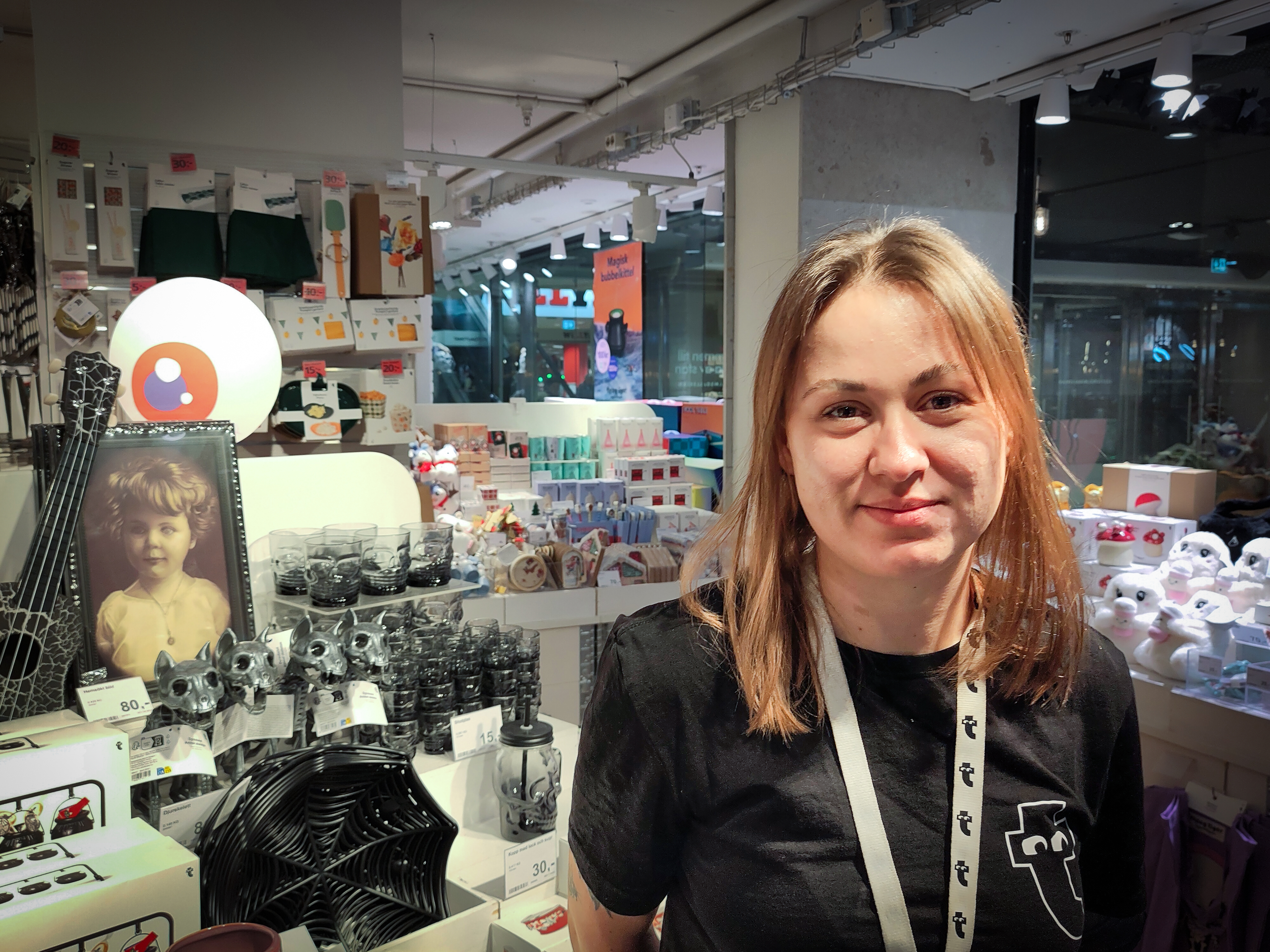 En kvinna med ett nyckelband står i en butiksgång med olika hushålls- och prydnadsföremål.