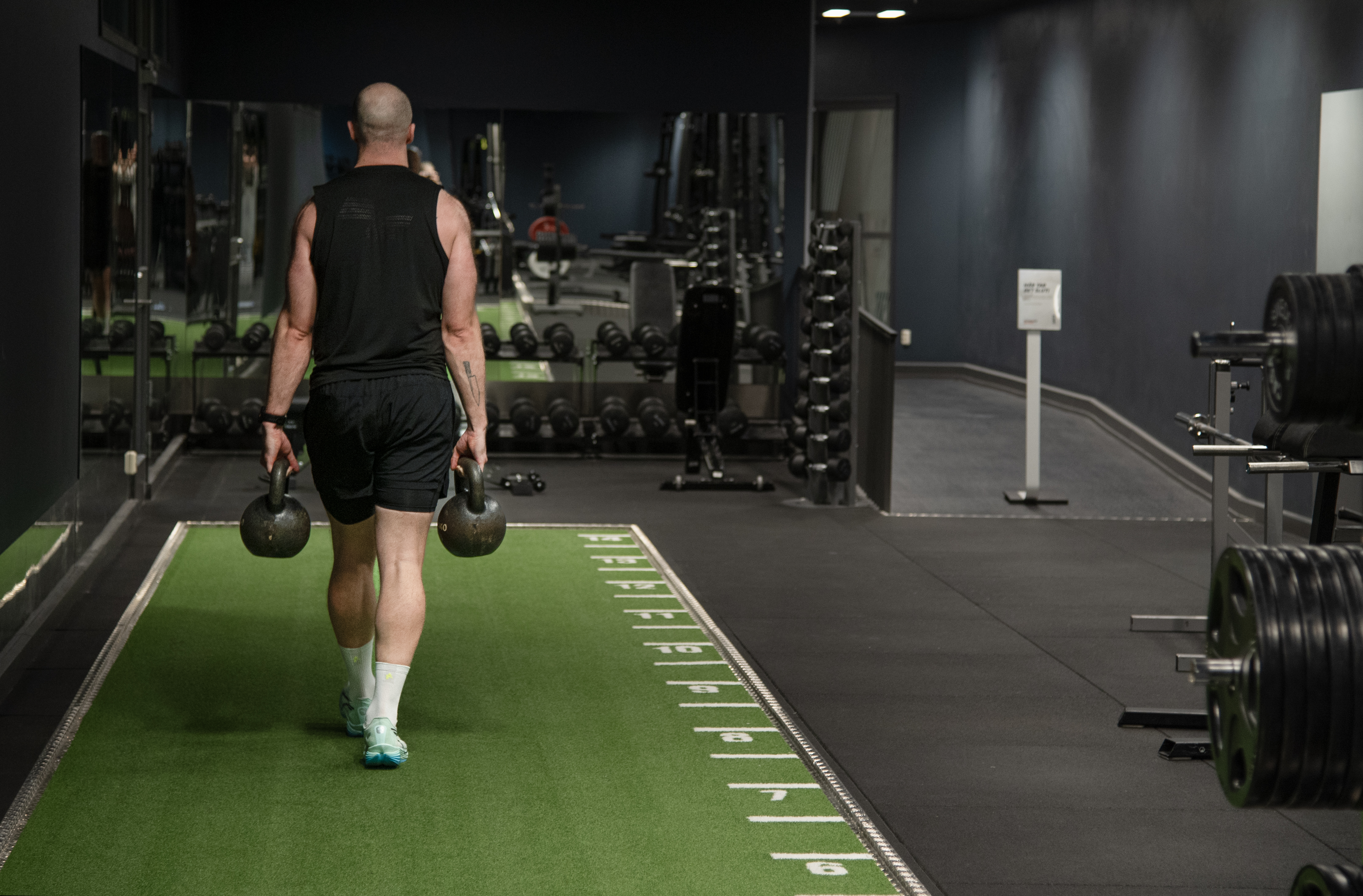 En man i träningskläder går på konstgräs inomhus och bär kettlebells i varje hand på ett gym med vikter och speglar.