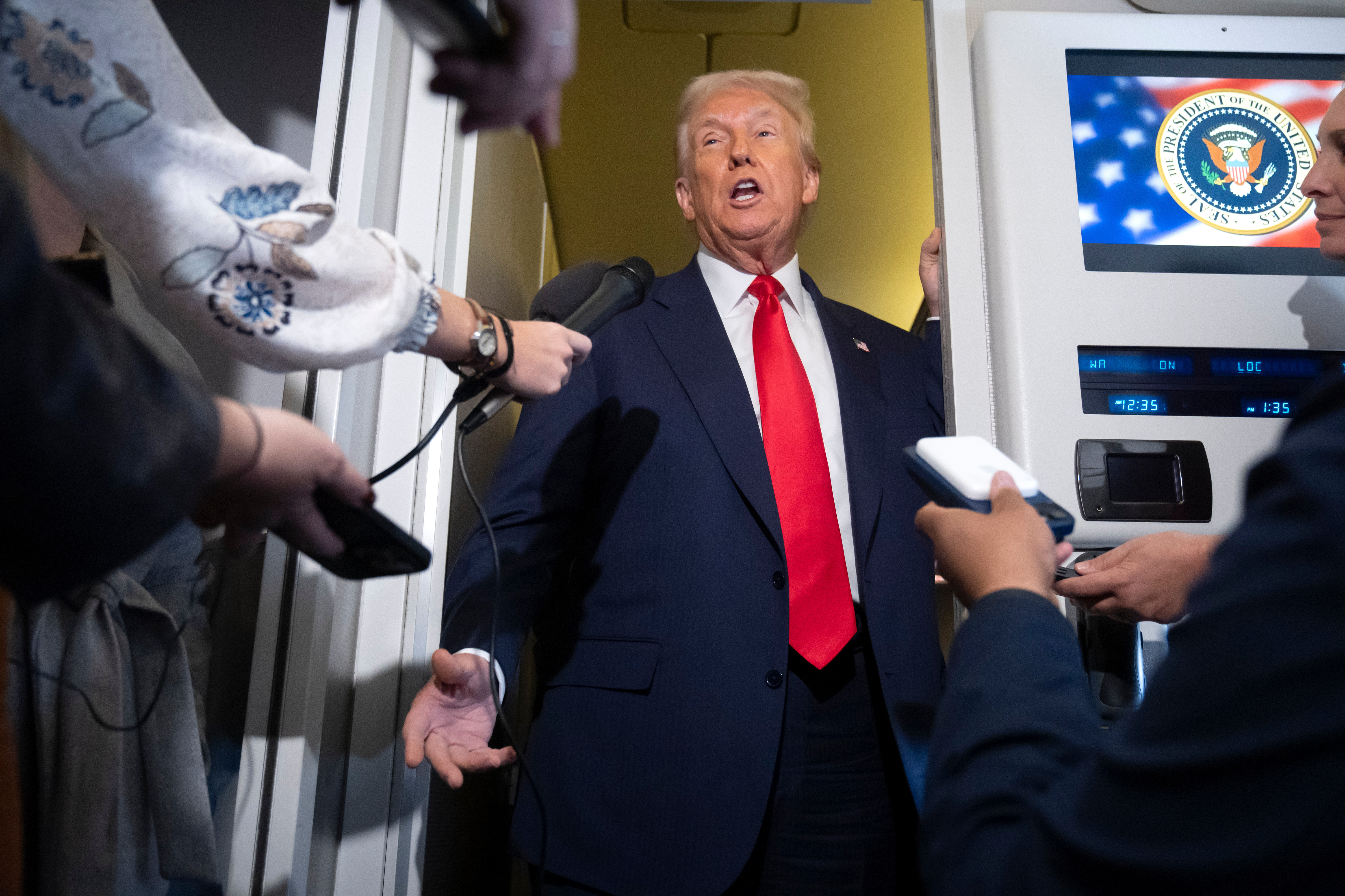 En man i kostym talar med reportrar som håller mikrofoner och inspelningsutrustning, med en skärm med en sigill i bakgrunden.