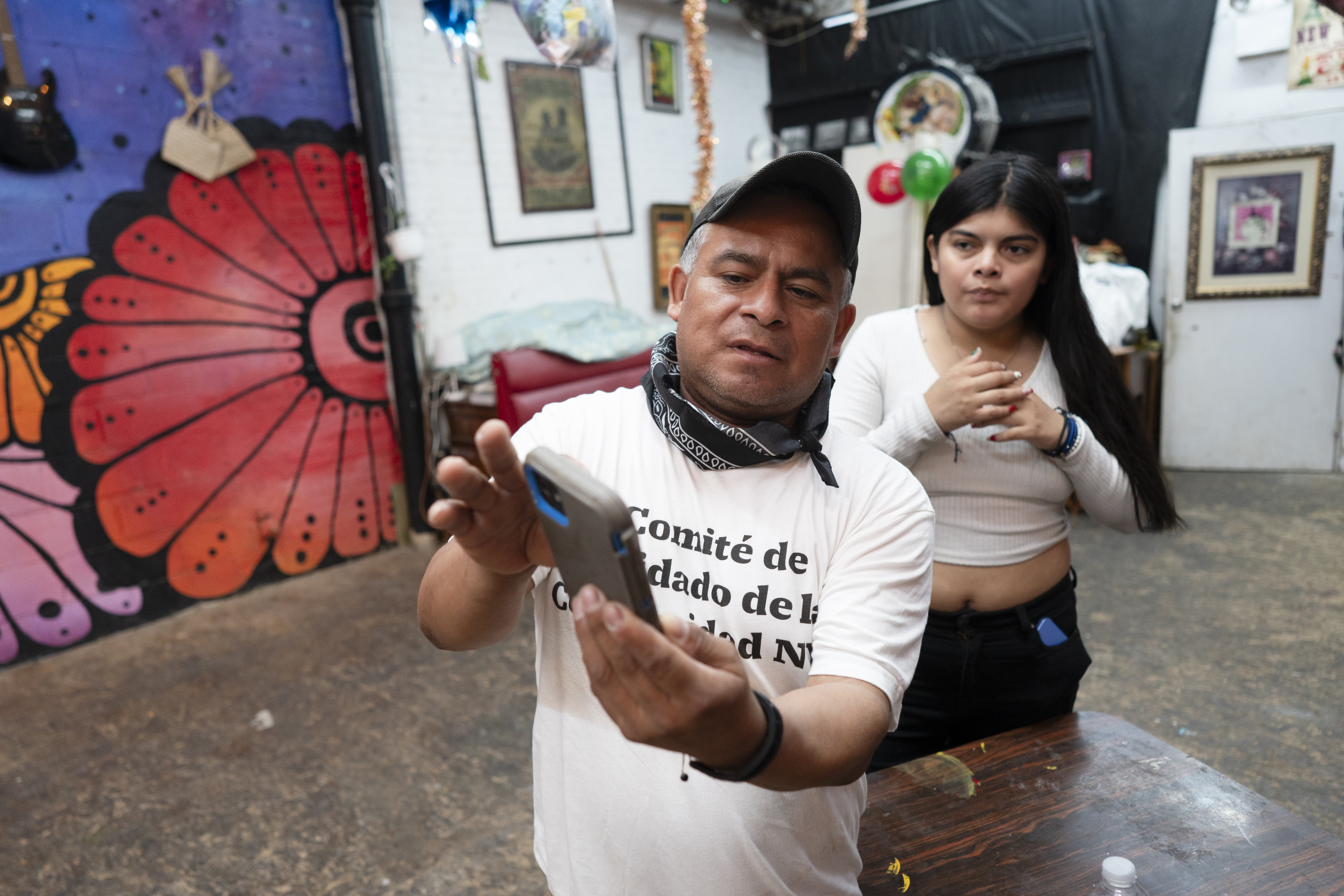 Crispin med vit t-shirt och keps tar en selfie med sin mobil, medan en Ana står bredvid i ett rum med konst och föremål.