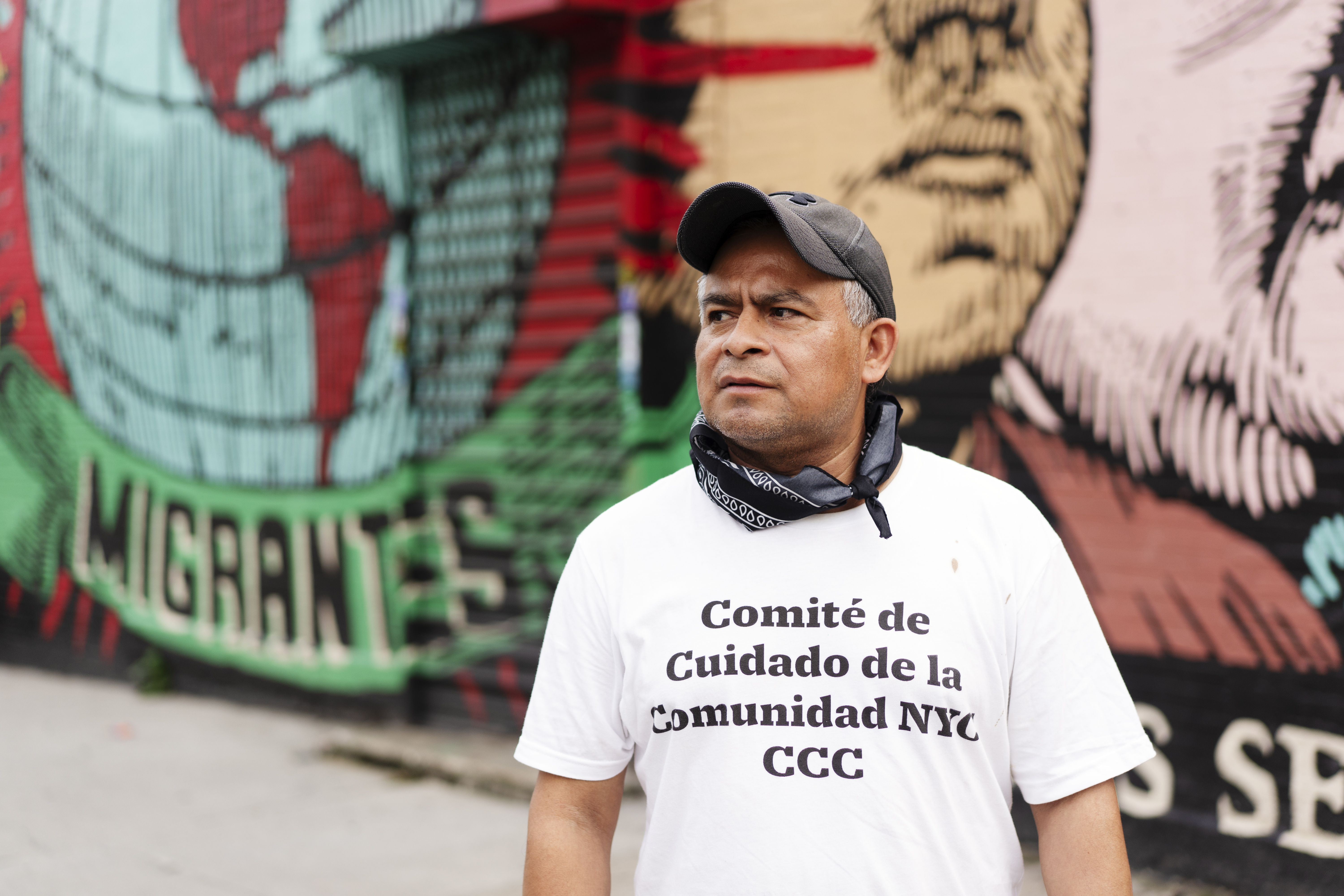 Crispin med en "Comité de Cuidado de la Comunidad NYC CCC"-tröja står framför en väggmålning där ordet "MIGRANTES" syns.
