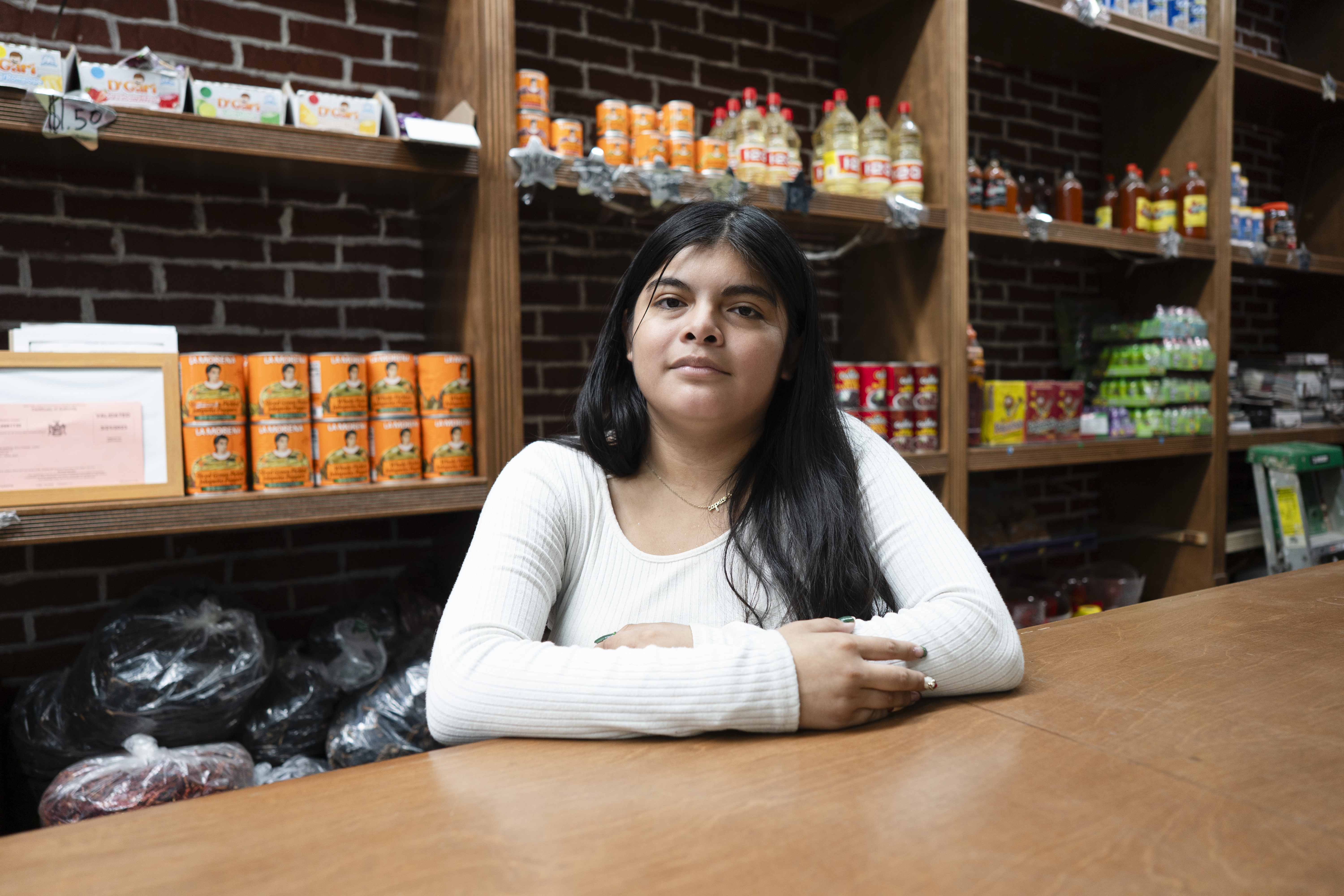 En kvinna i vit skjorta sitter vid en disk i en butik med hyllor med matvaror och andra varor bakom sig.