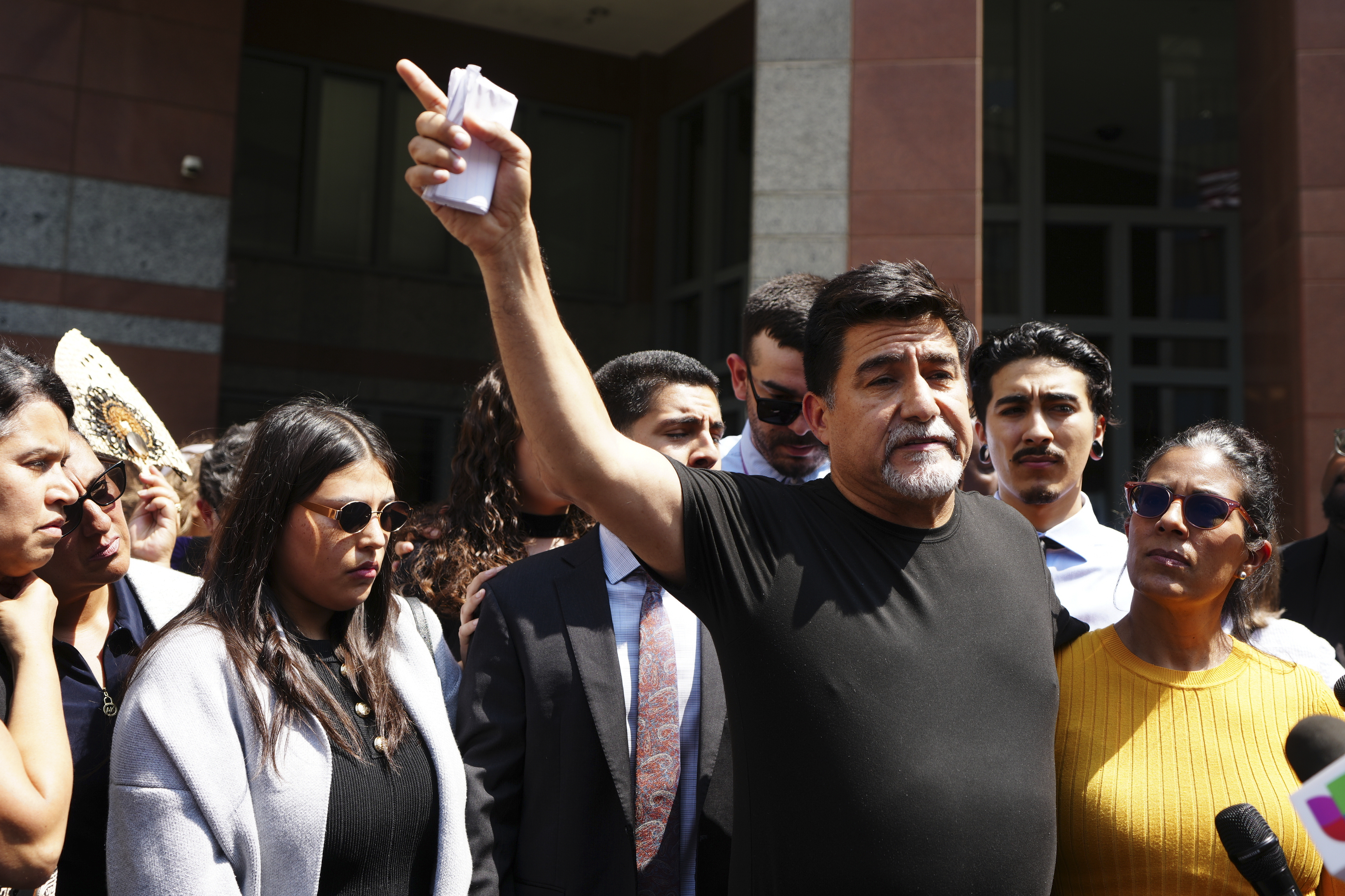 David Huerta i svart skjorta talar till en folkmassa utomhus, höjer armen och håller papper. Folk står runt honom och lyssnar.