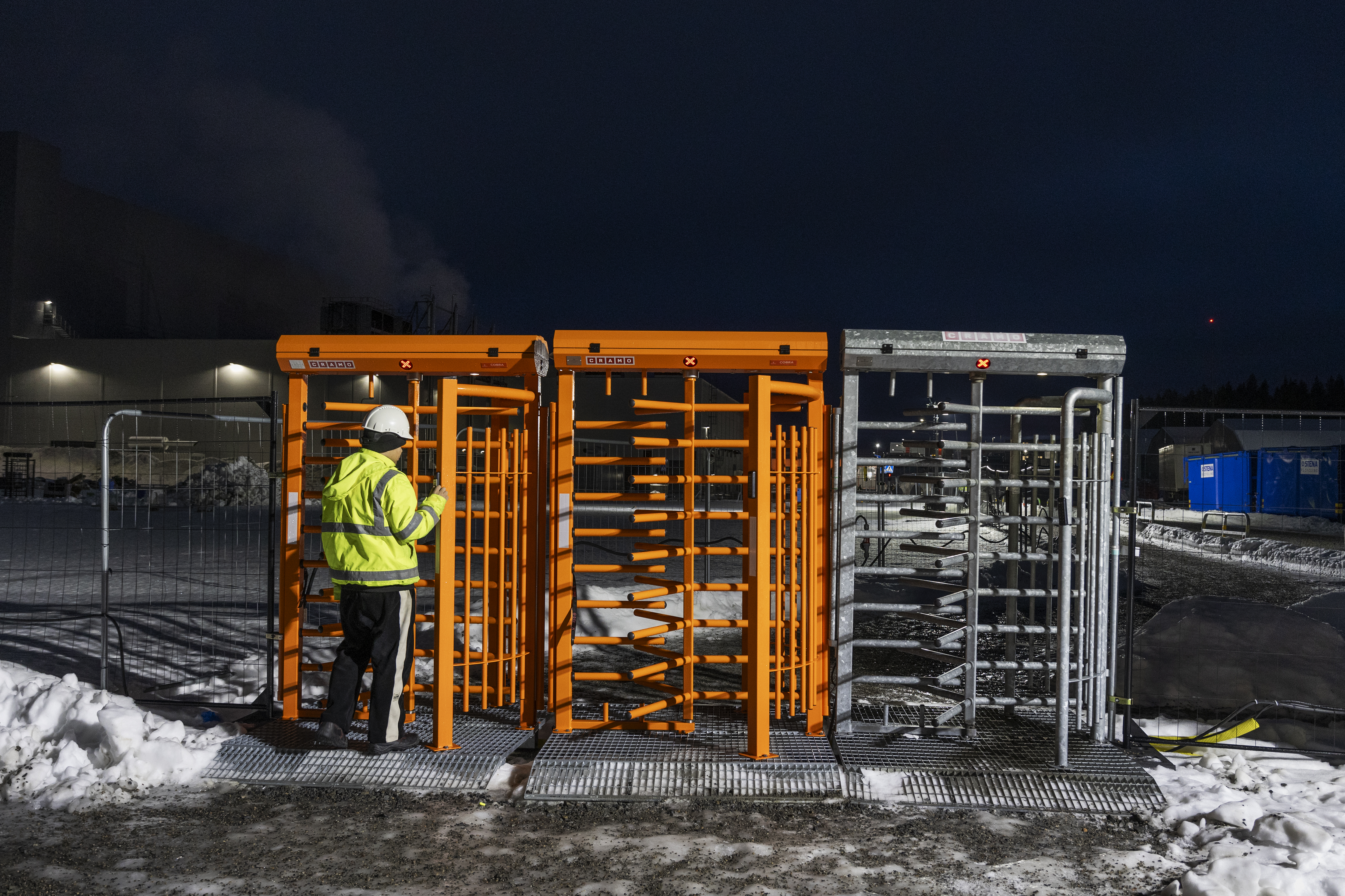 En arbetare med reflexjacka och hjälm står vid orange säkerhetsspärrar på en snötäckt industriplats på natten.