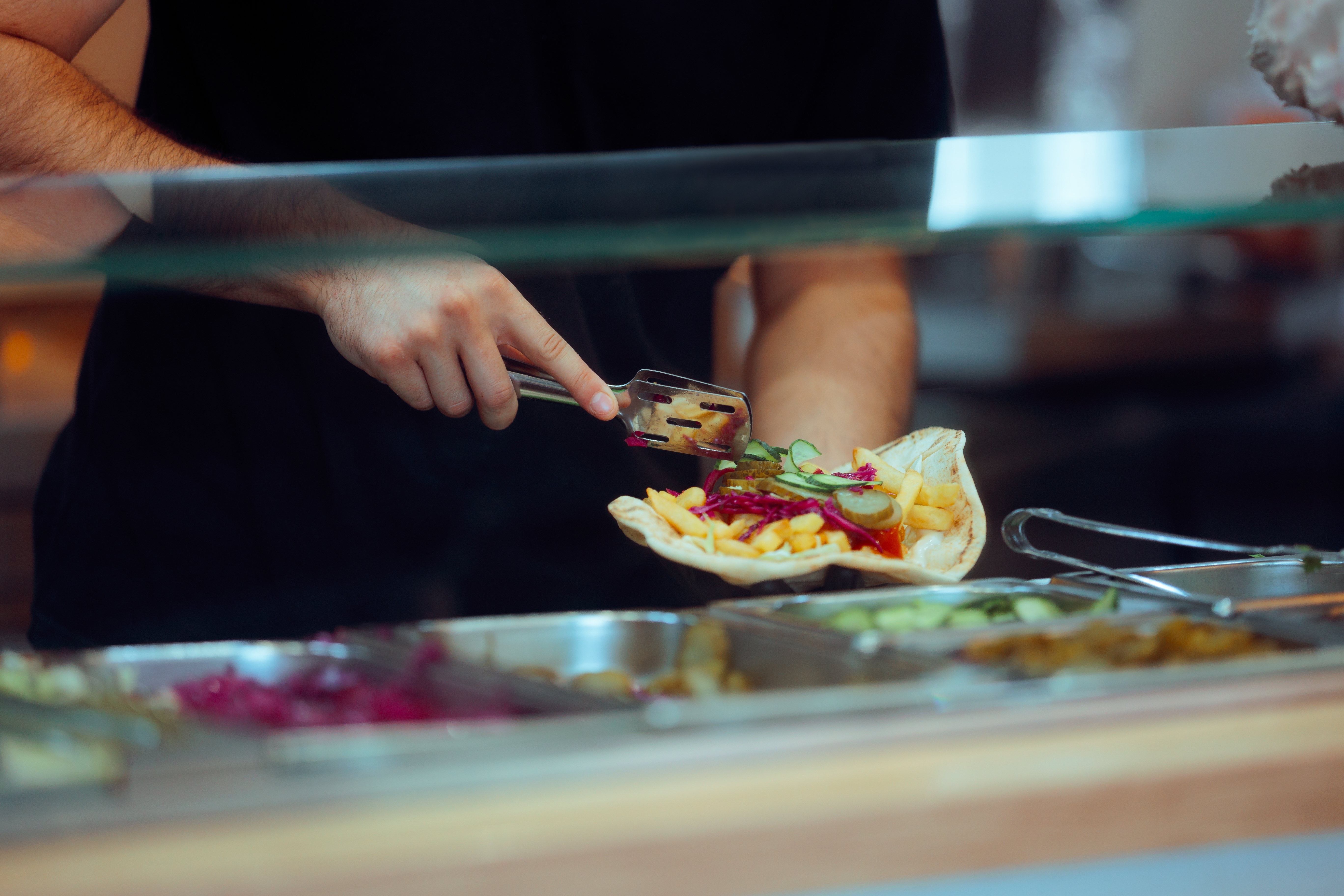 En person gör en wrap med grönsaker och pommes bakom en glasskiva vid en serveringsdisk.