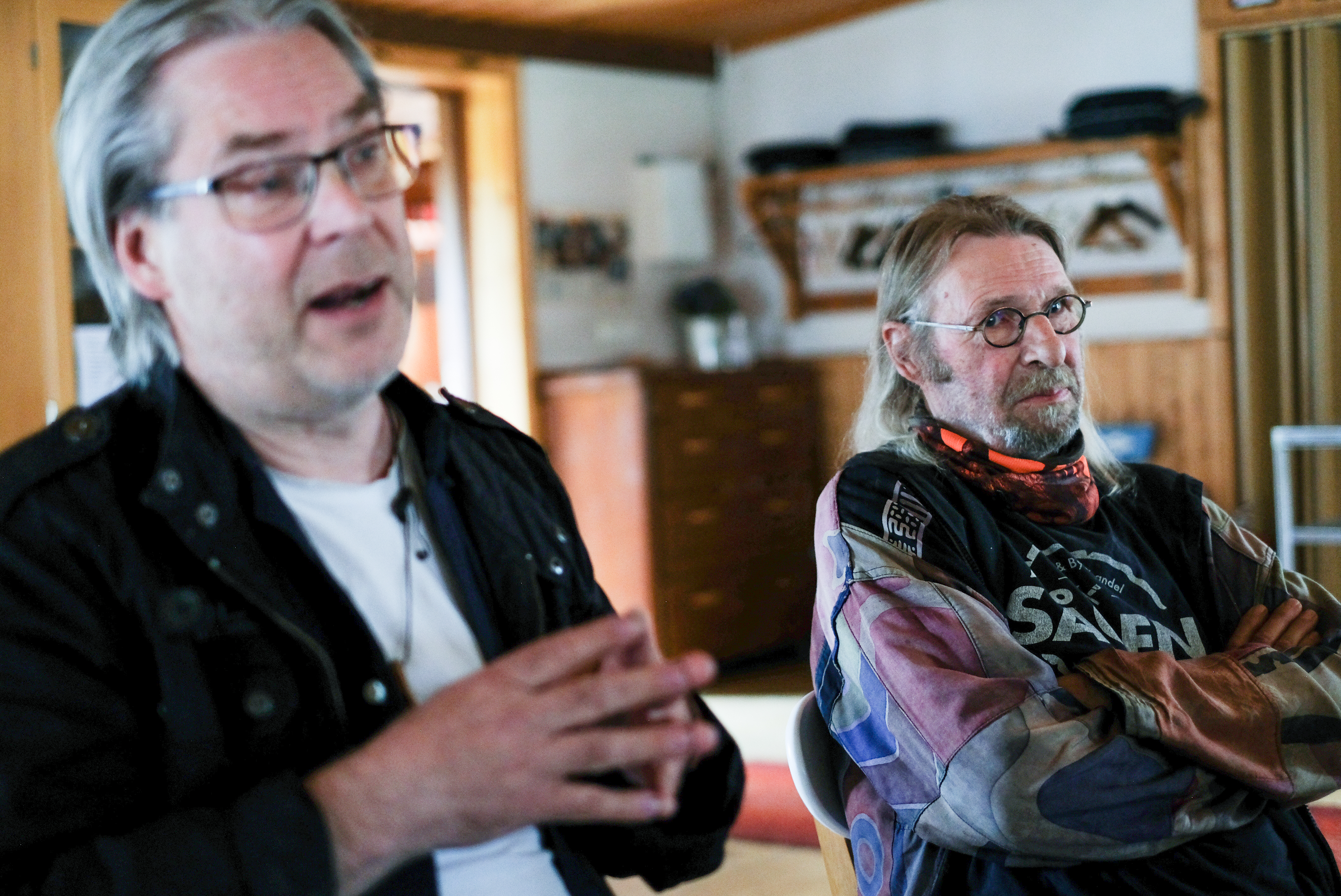 Mats Sidmalm och Ulrik Lidström sitter inomhus; Mats pratar och gestikulerar, Ulrik lyssnar med armarna i kors och har glasögon och jacka.