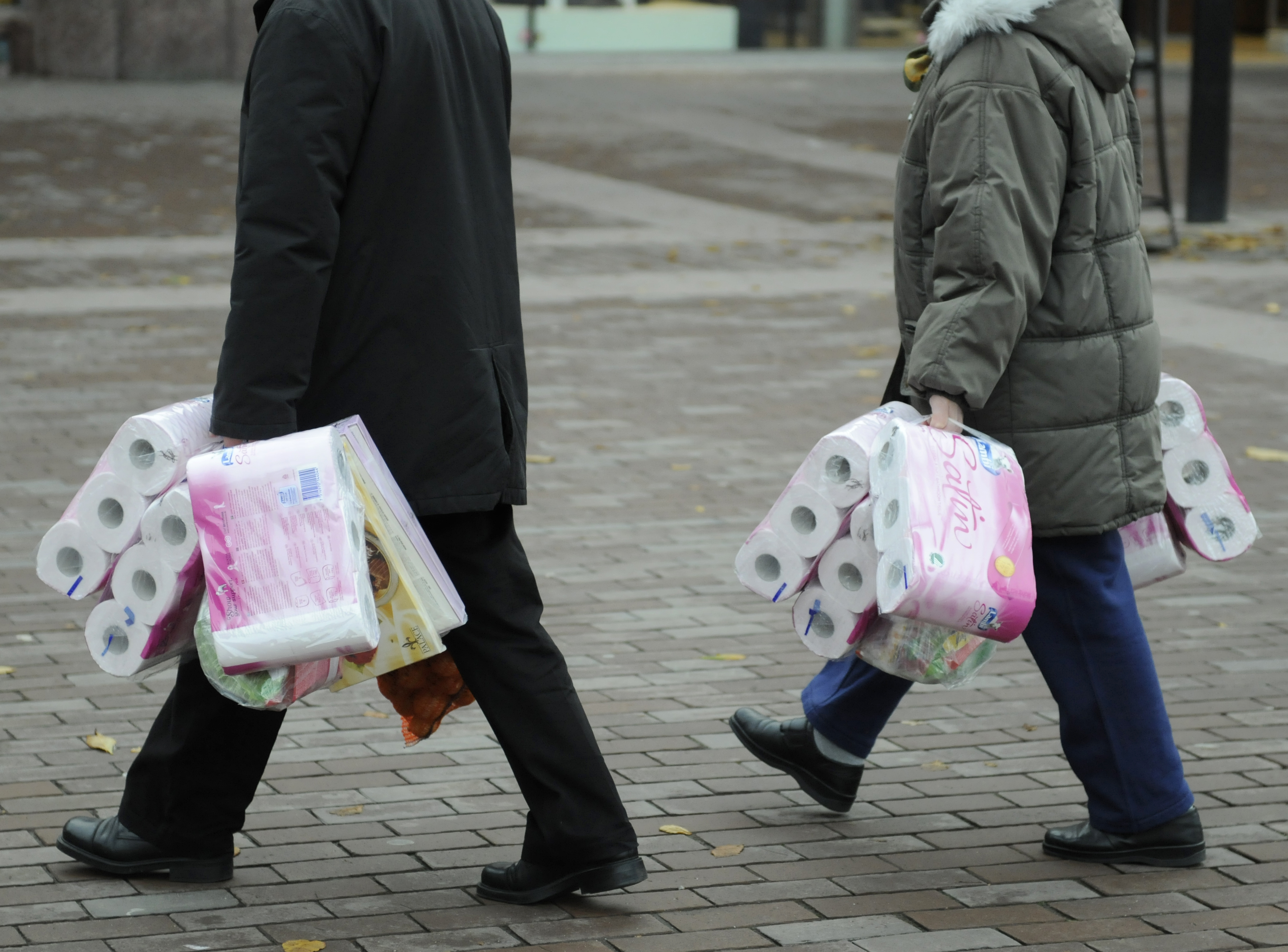 De går sida vid sida på trottoaren och bär på flera paket toalettpapper och pappershanddukar.