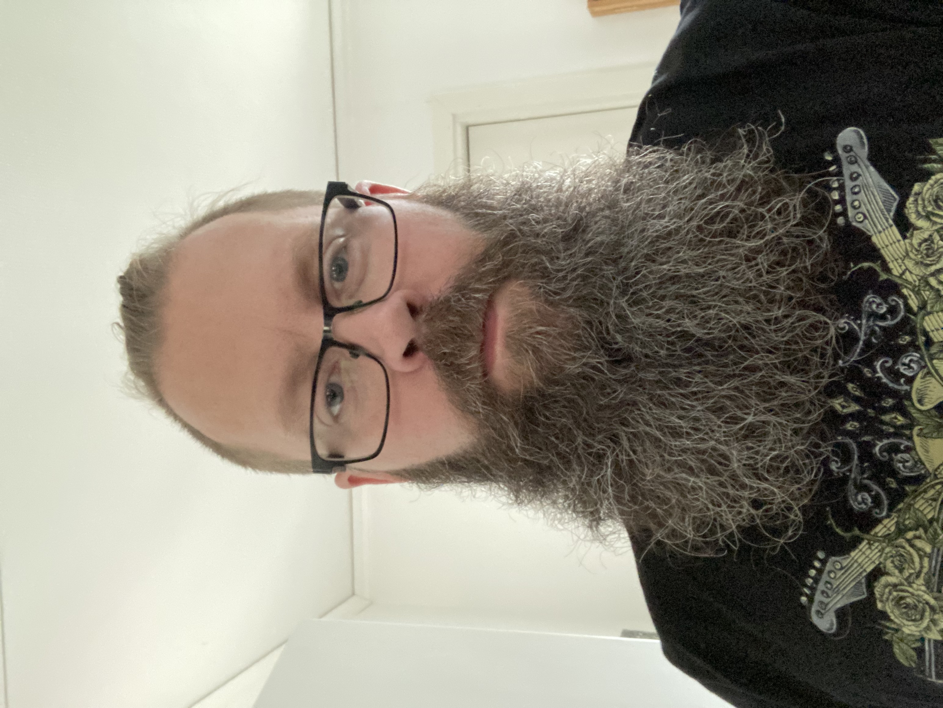 En person med glasögon och långt skägg står inomhus och tittar mot kameran med neutral min.