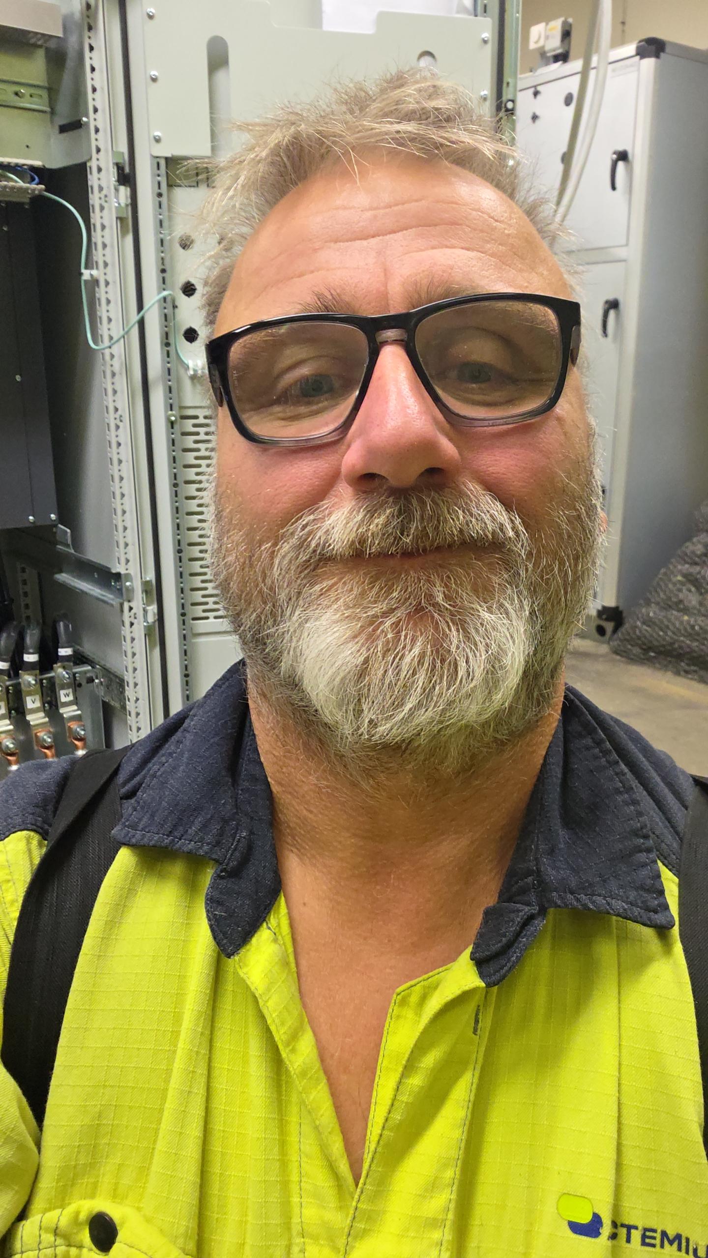 En man med glasögon och en gul varseltröja står framför elektrisk utrustning och tar en selfie.