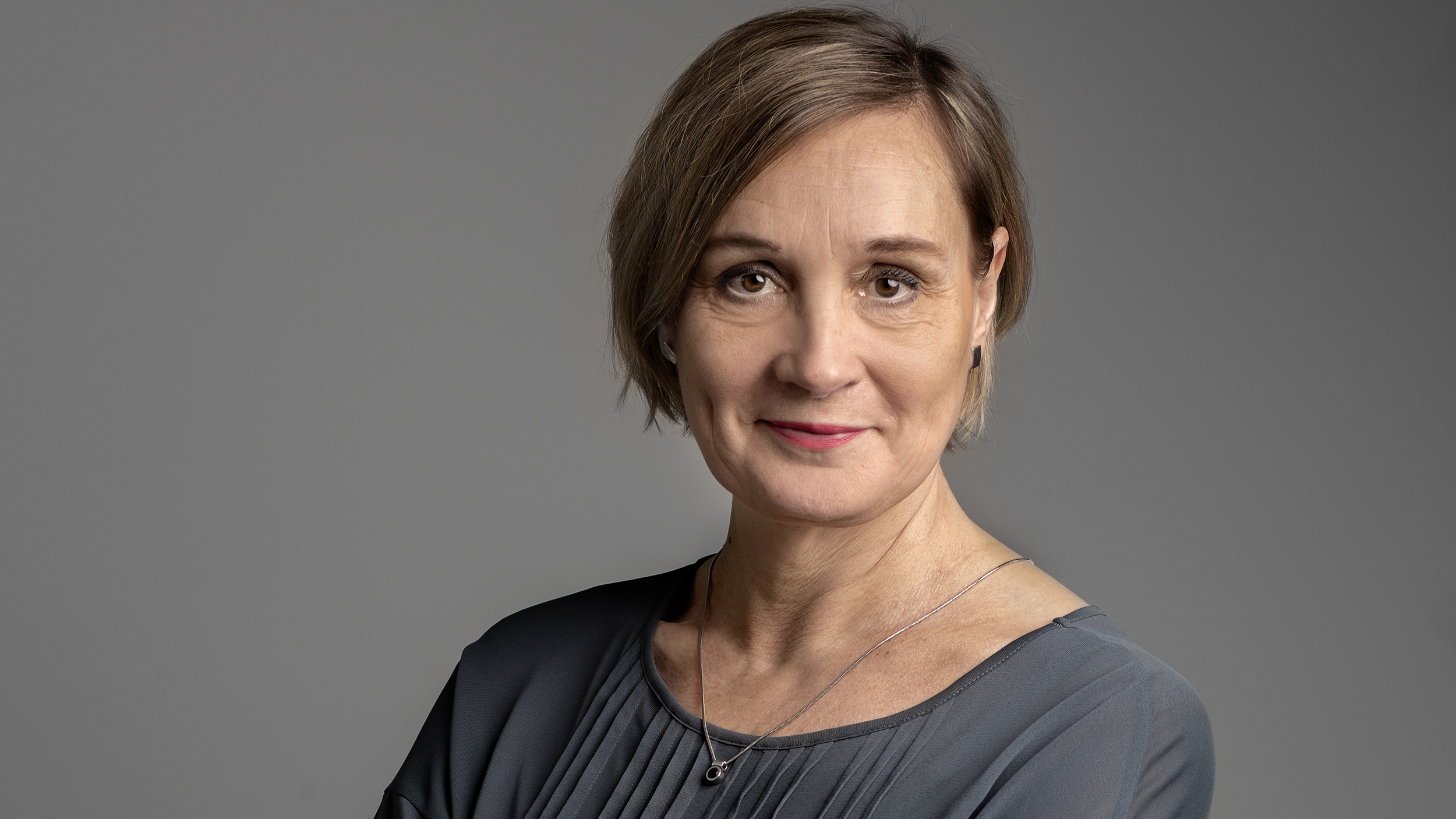 En kvinna med kort ljusbrunt hår och grå blus står mot en grå bakgrund och tittar in i kameran med neutral min.