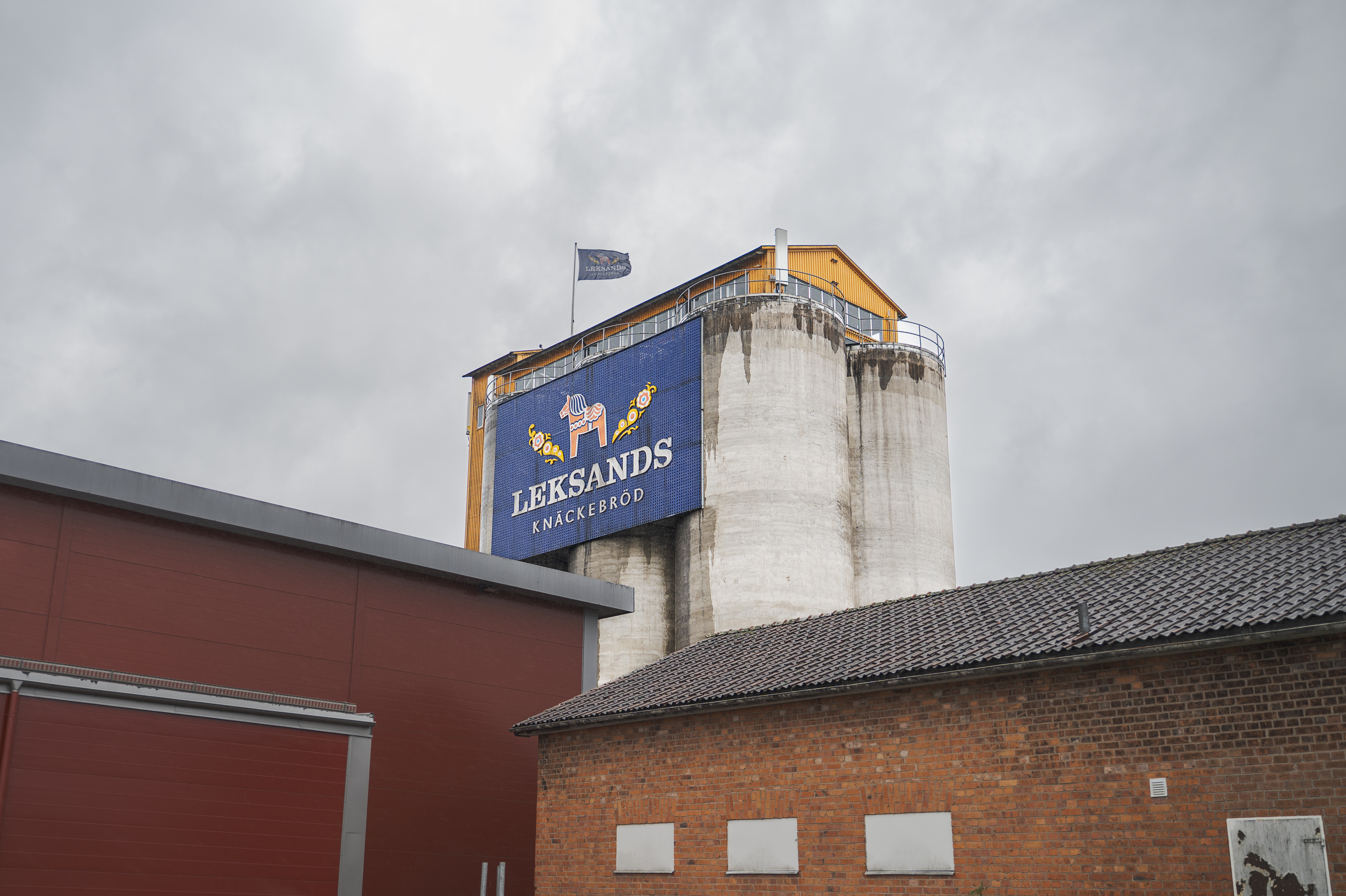 En silo i betong med en skylt och flagga där det står "Leksands Knäckebröd" står bakom röda och bruna industribyggnader.