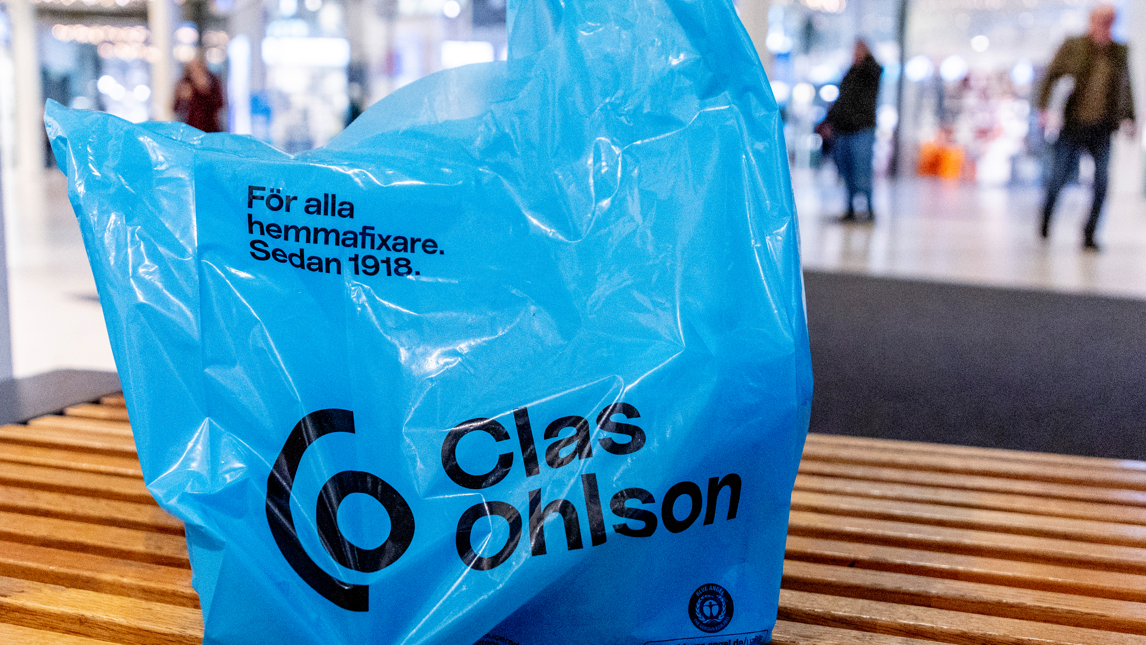En plastpåse från Clas Ohlson står på en bänk i ett köpcentrum. I bakgrunden syns människor, men de är suddiga.