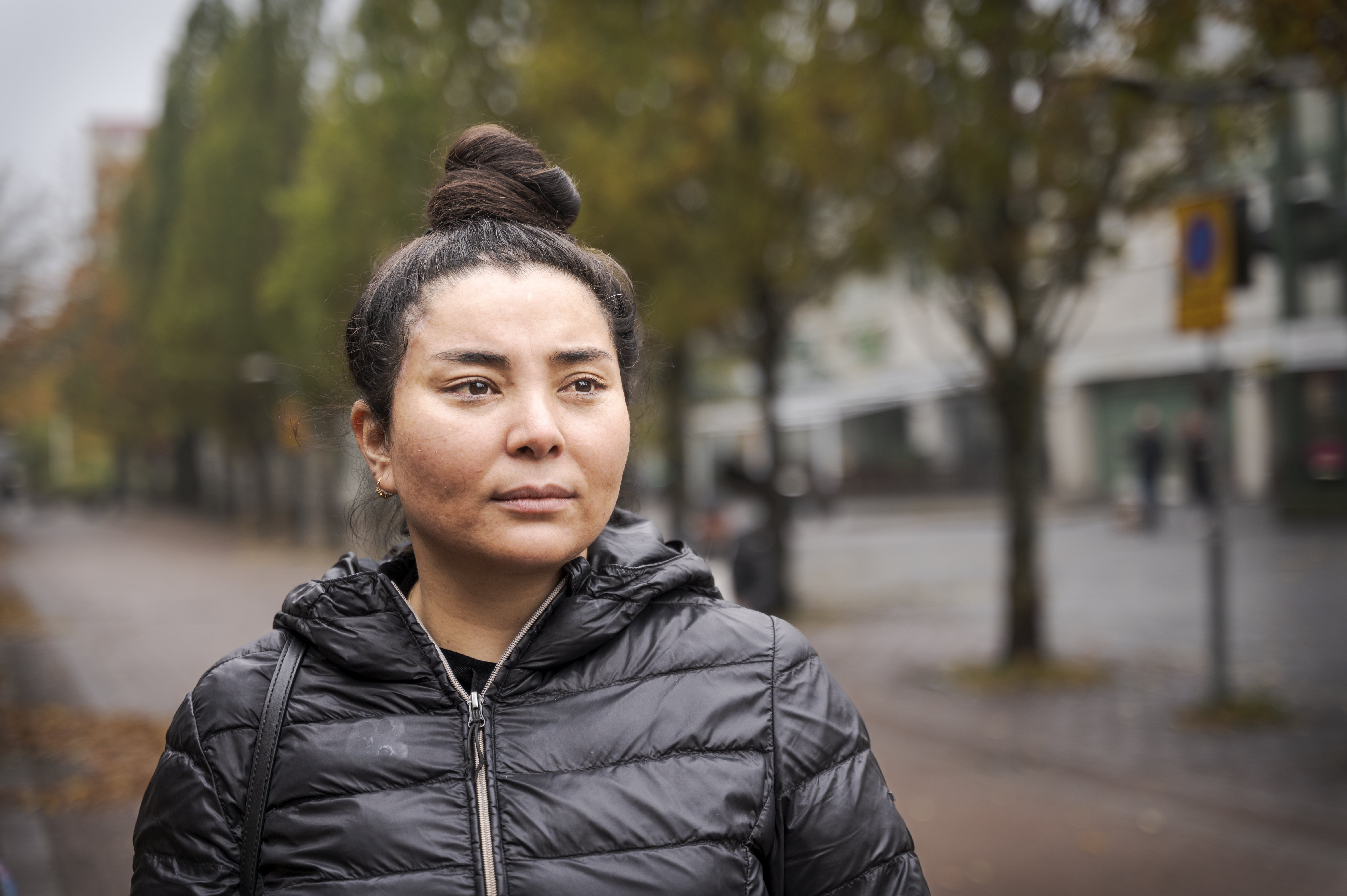 Rihana Ibrahimi med mörkt hår i en knut bär en svart dunjacka och står utomhus på en gata med träd och suddiga byggnader.