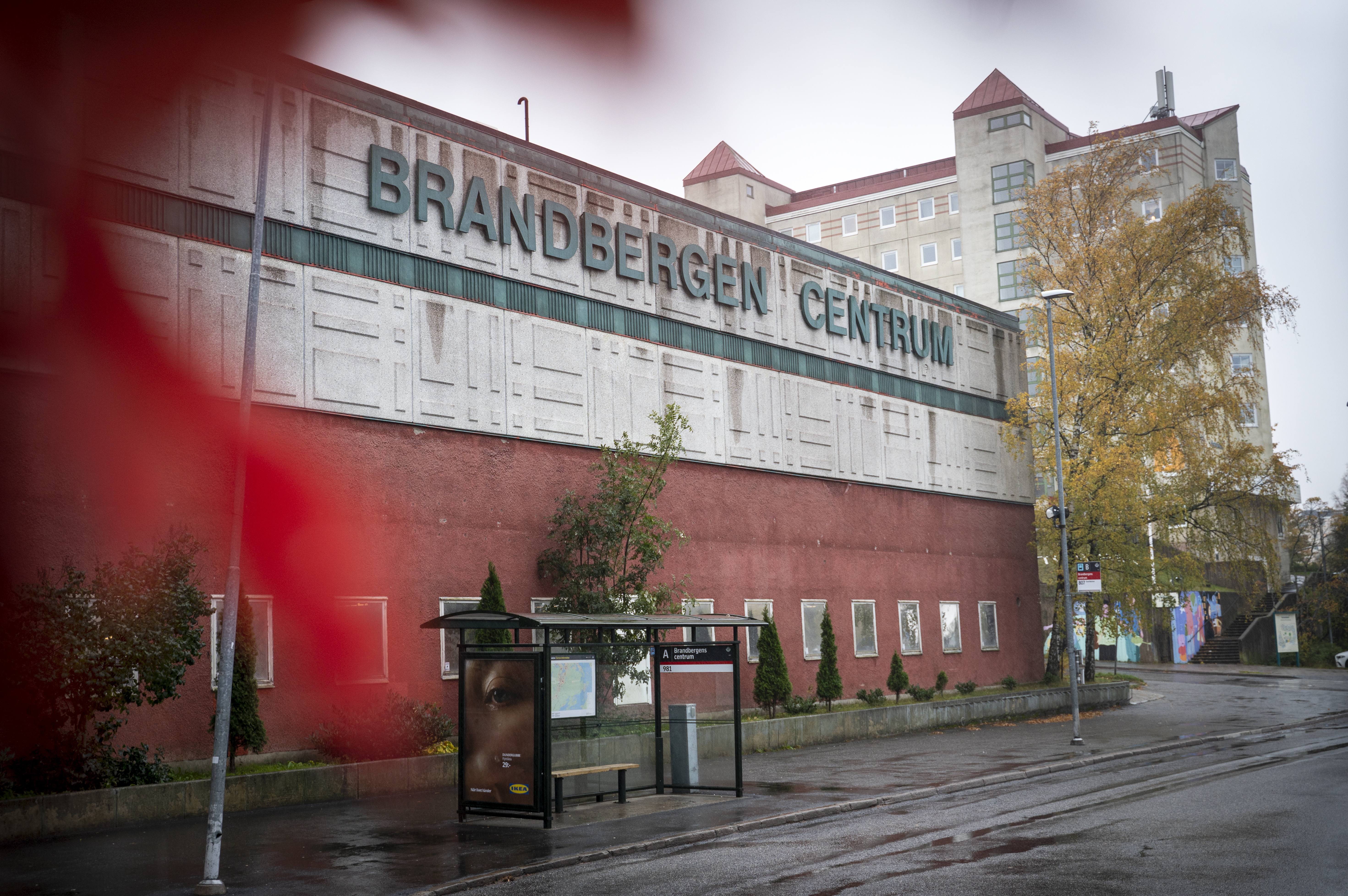 En gatubild av Brandbergen Centrum med en busshållplats framför. Några träd och en affisch syns nära hållplatsen.