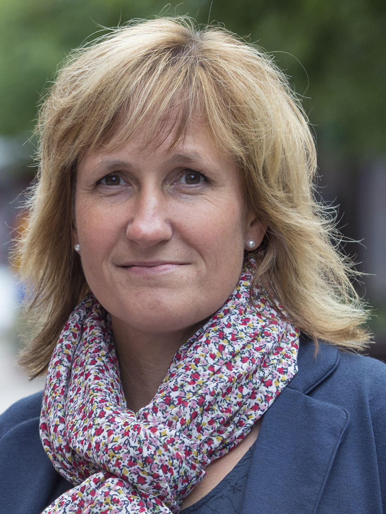 Anna Hed med blont hår, en blommig scarf och mörk jacka står utomhus och tittar på kameran med neutral min.