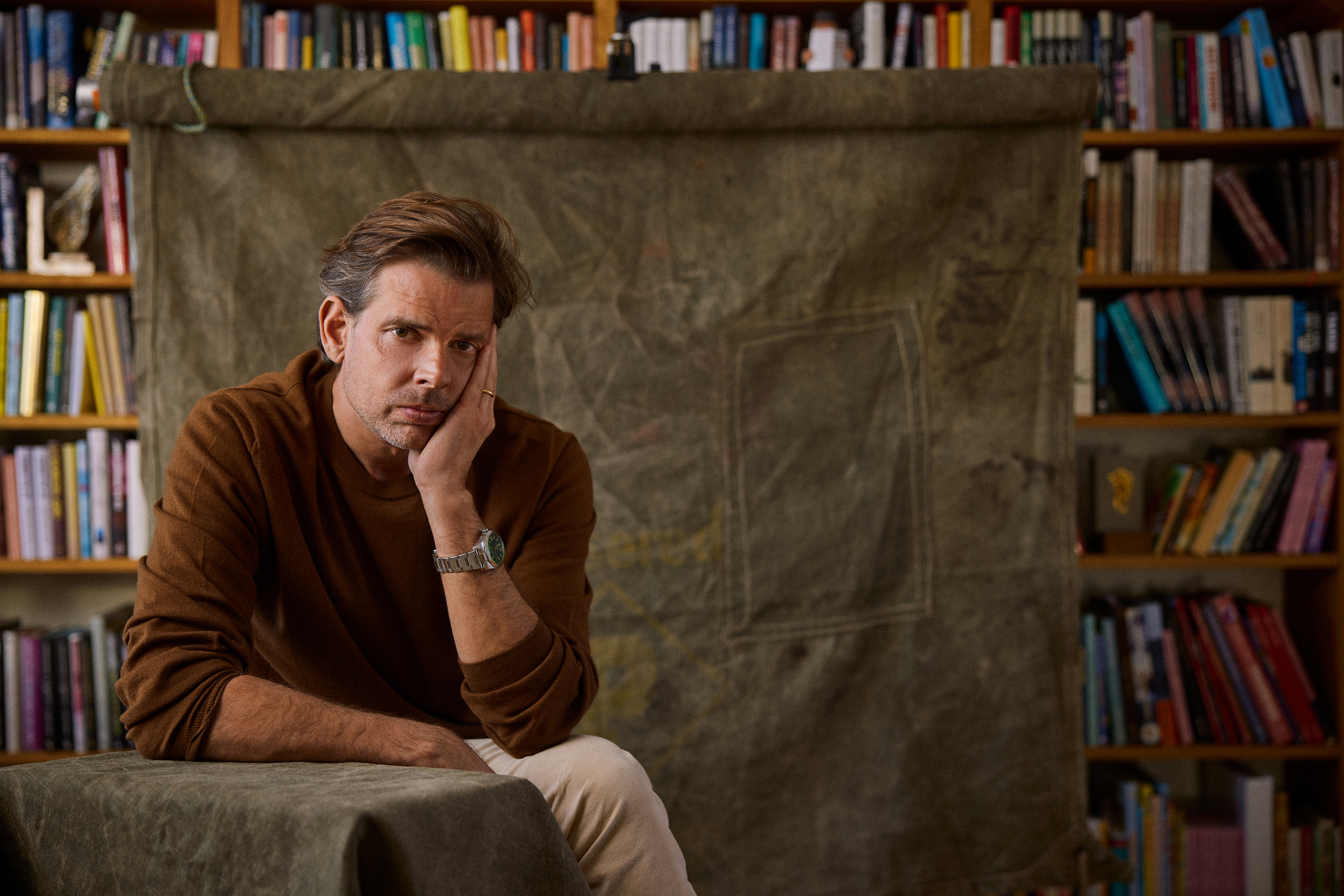 Författaren Alex Schulman med en brun tröja sitter med huvudet mot handen framför en täckt duk och bokhyllor fyllda med böcker.