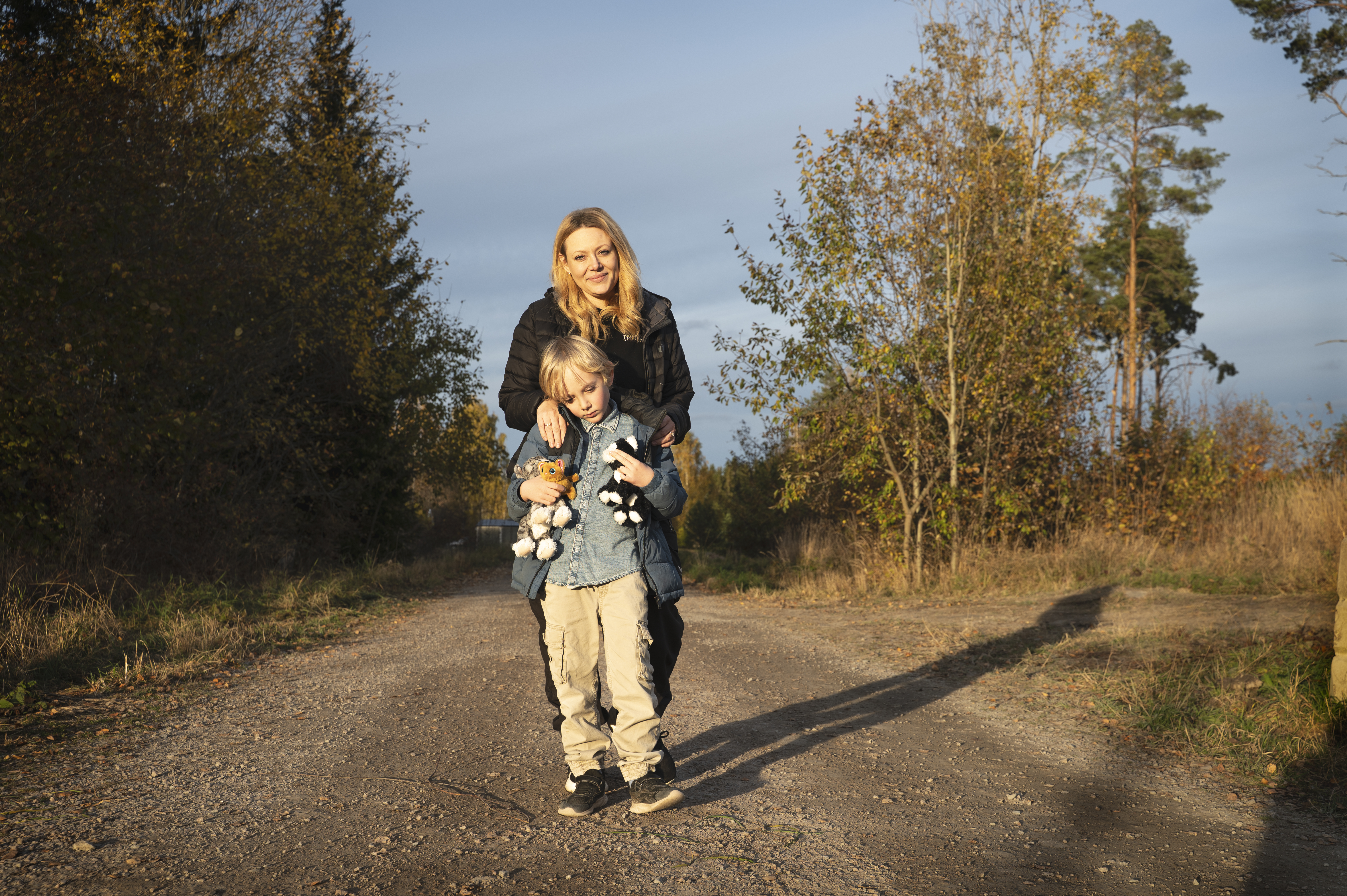 En kvinna står på en grusväg med armarna runt en pojke som håller gosedjur, omgiven av träd under en klar himmel.