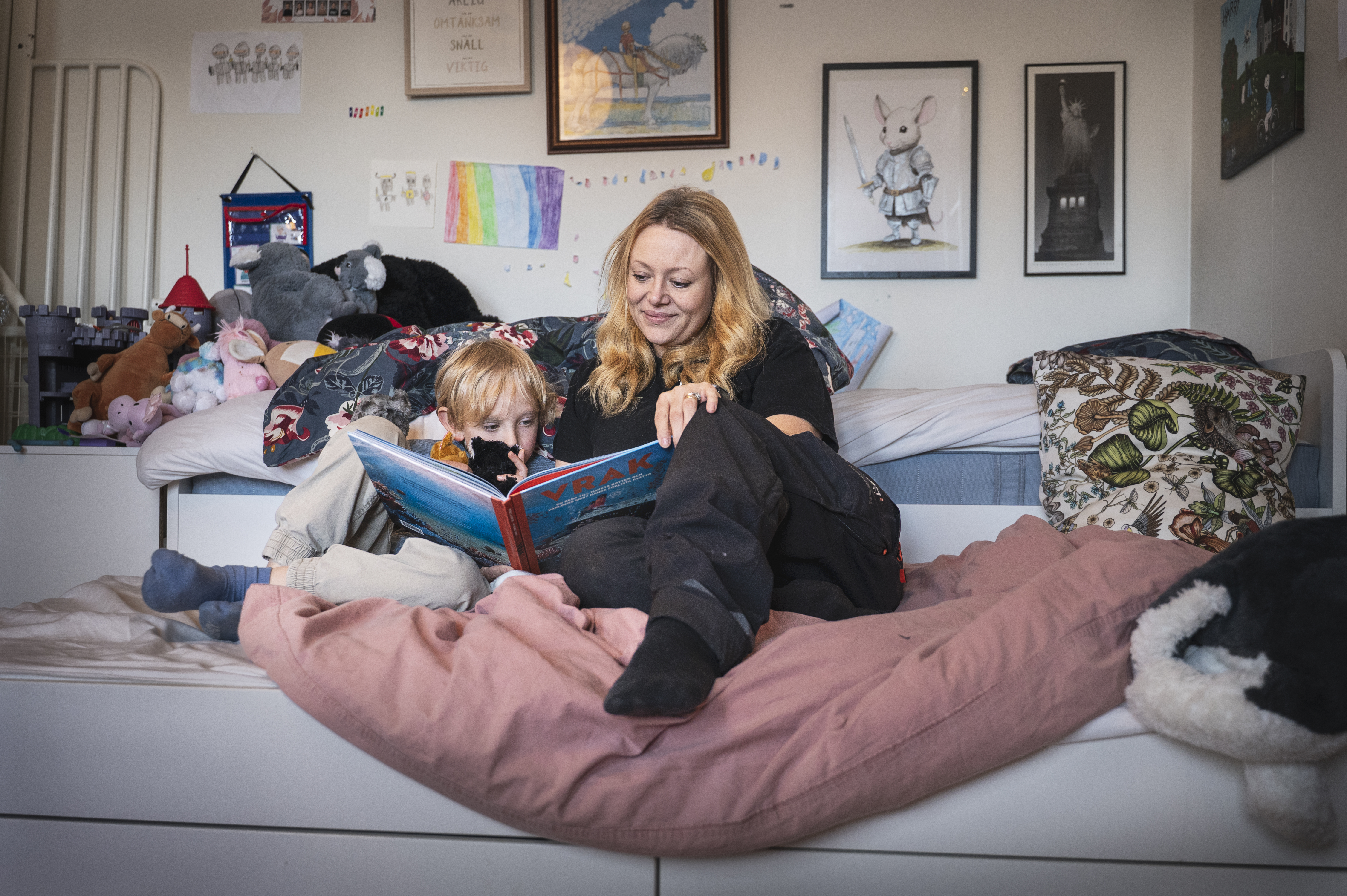 En vuxen och ett barn sitter på en säng och läser en bok i ett sovrum med leksaker, konst och teckningar på väggen.