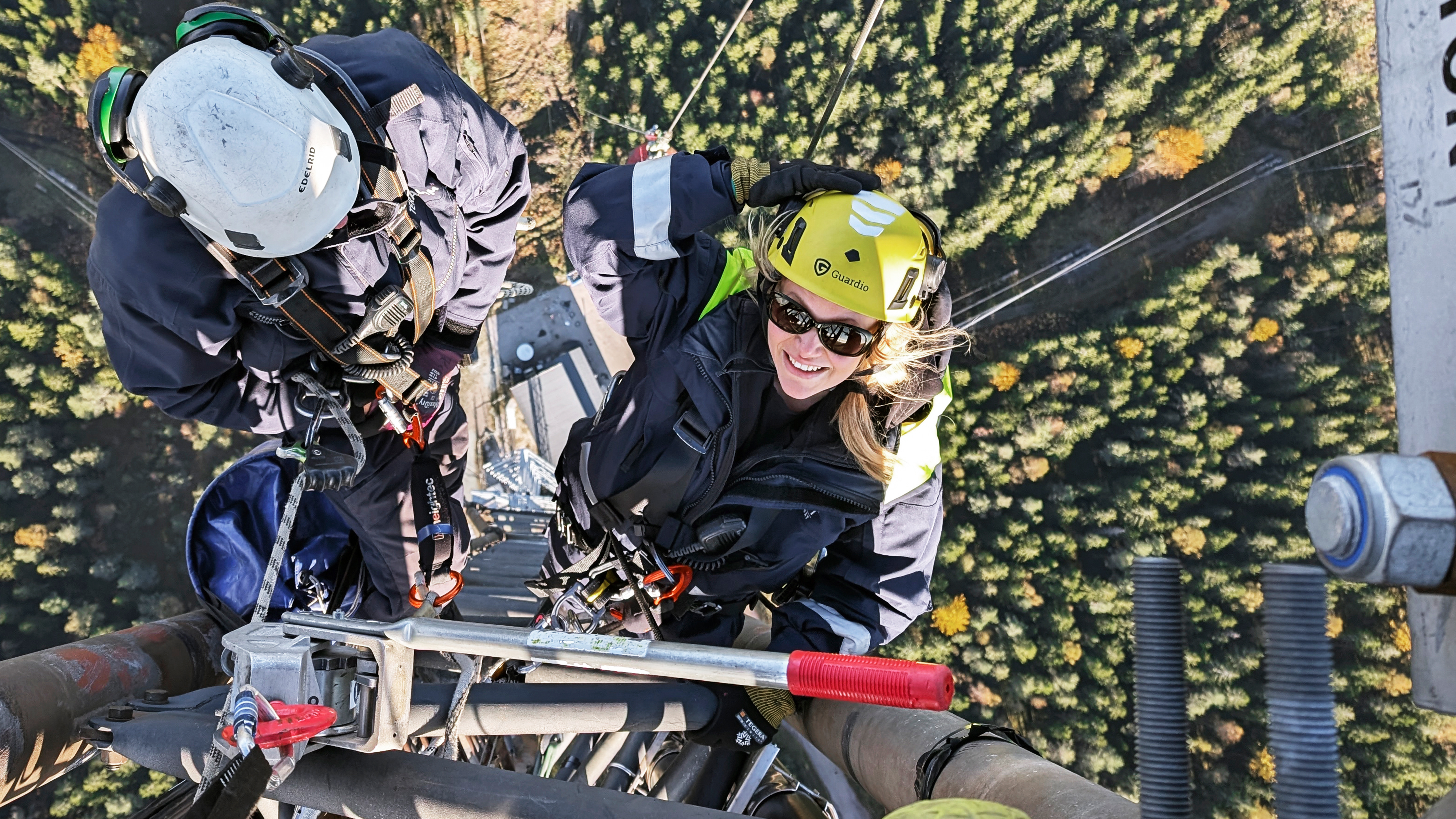 Två arbetare med skyddsutrustning och hjälmar utför underhåll på en telekommunikationstorn ovanförför ett skogsområde.