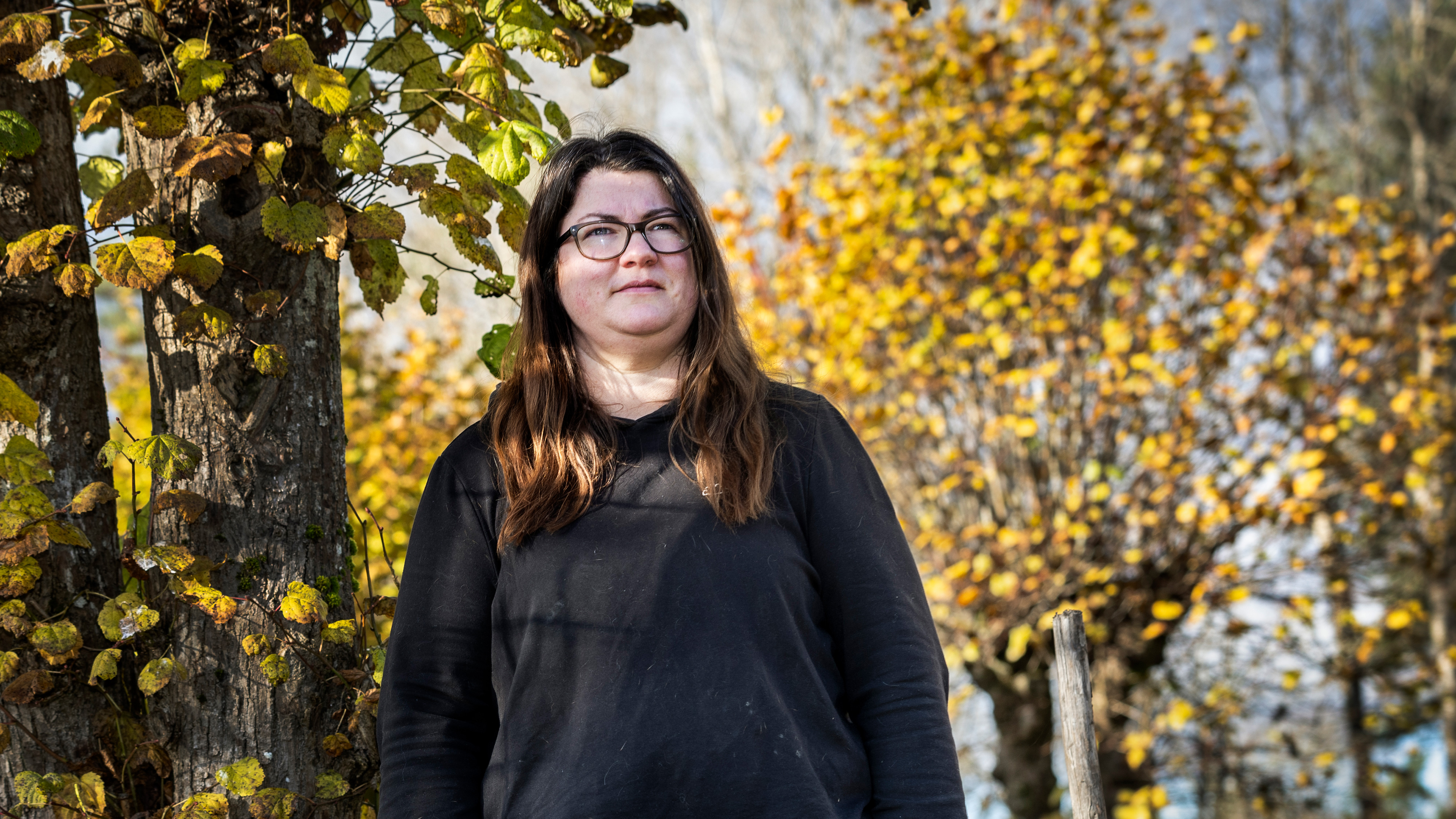 En person med långt brunt hår och glasögon står utomhus nära träd med gula höstlöv.