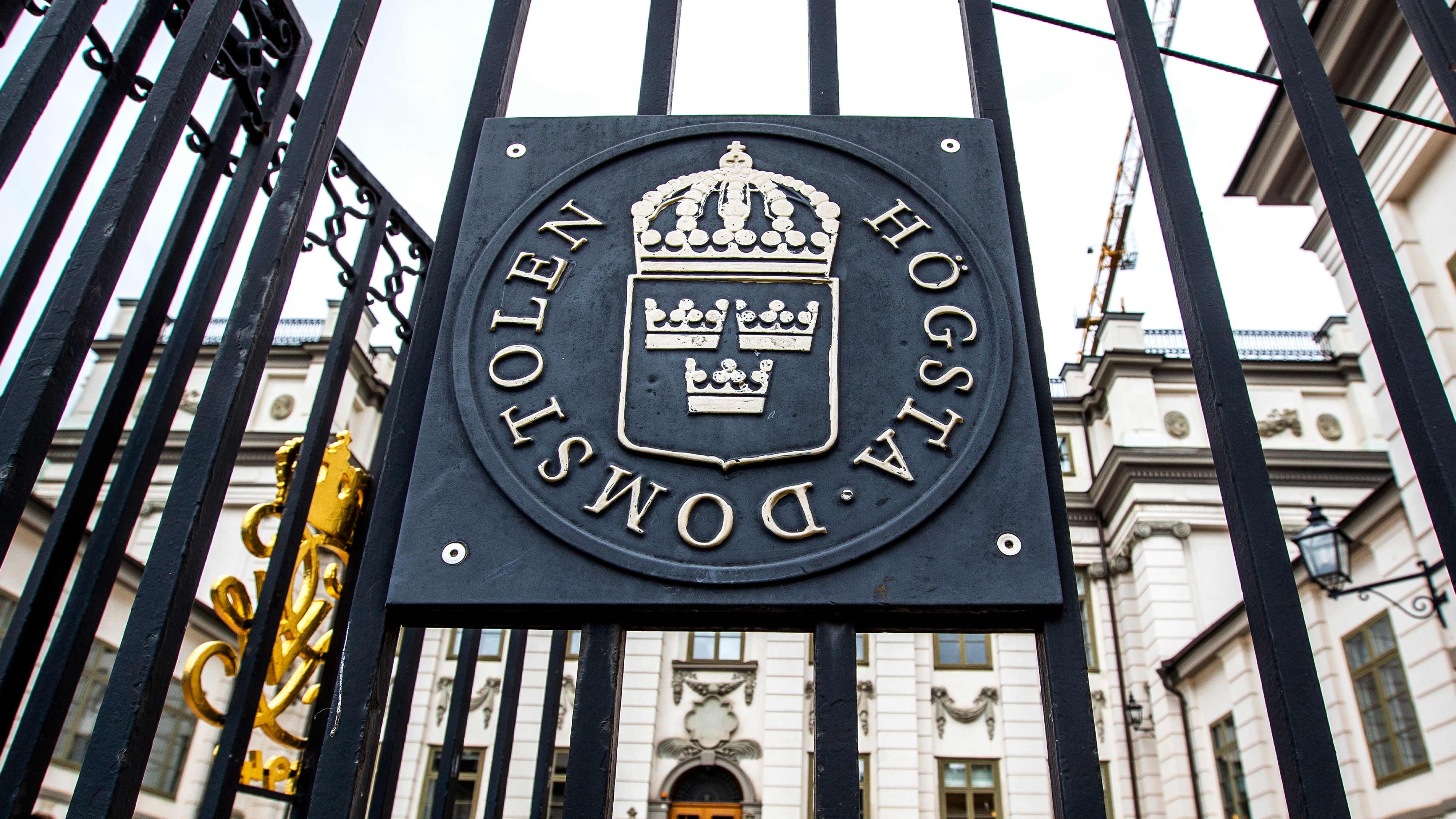 En metallskylt på en grind visar emblemet och texten Högsta domstolen. Byggnaden syns i bakgrunden.