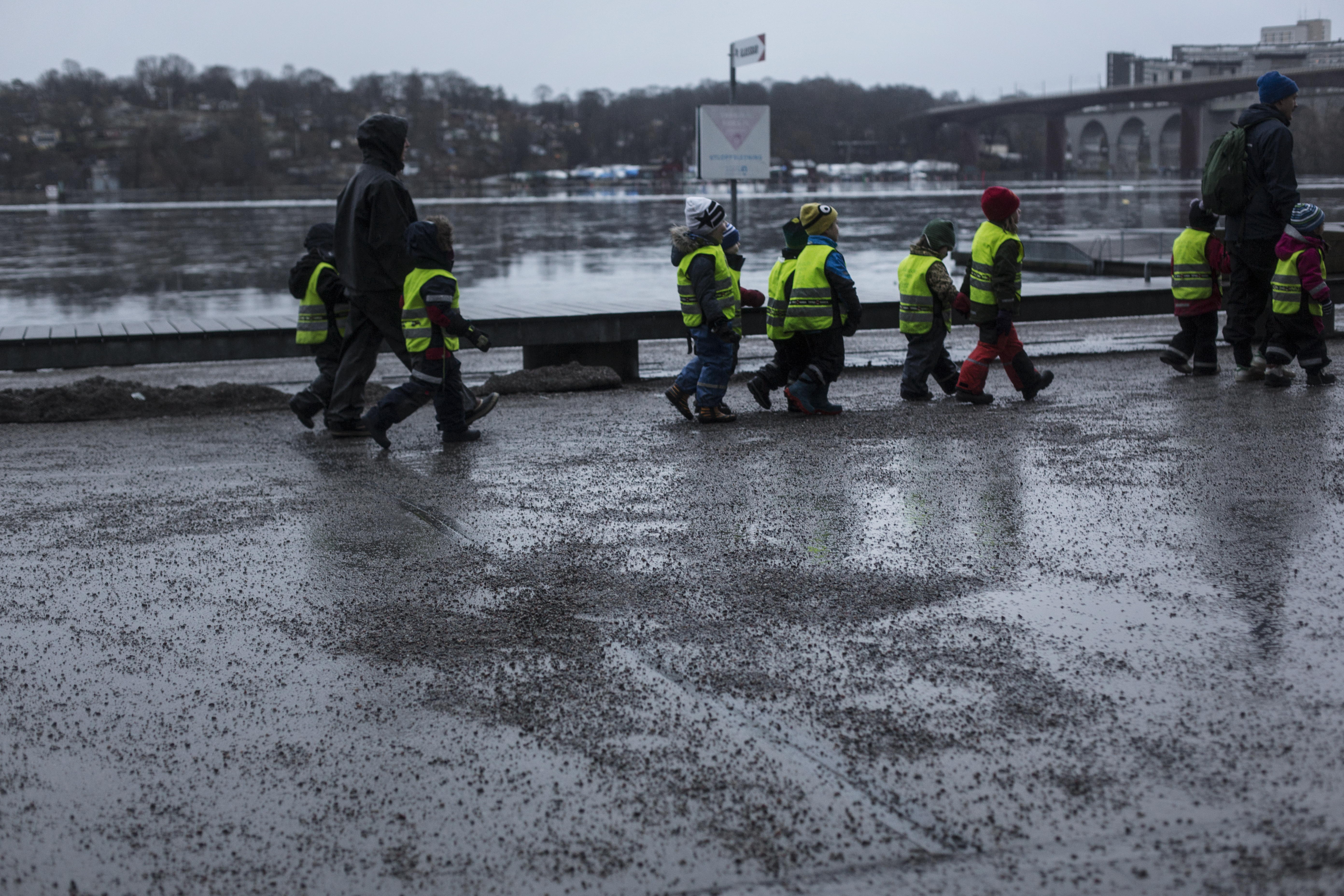 En grupp barn med reflexvästar går i rad längs en gångväg vid en flod, tillsammans med två vuxna under en molnig dag.