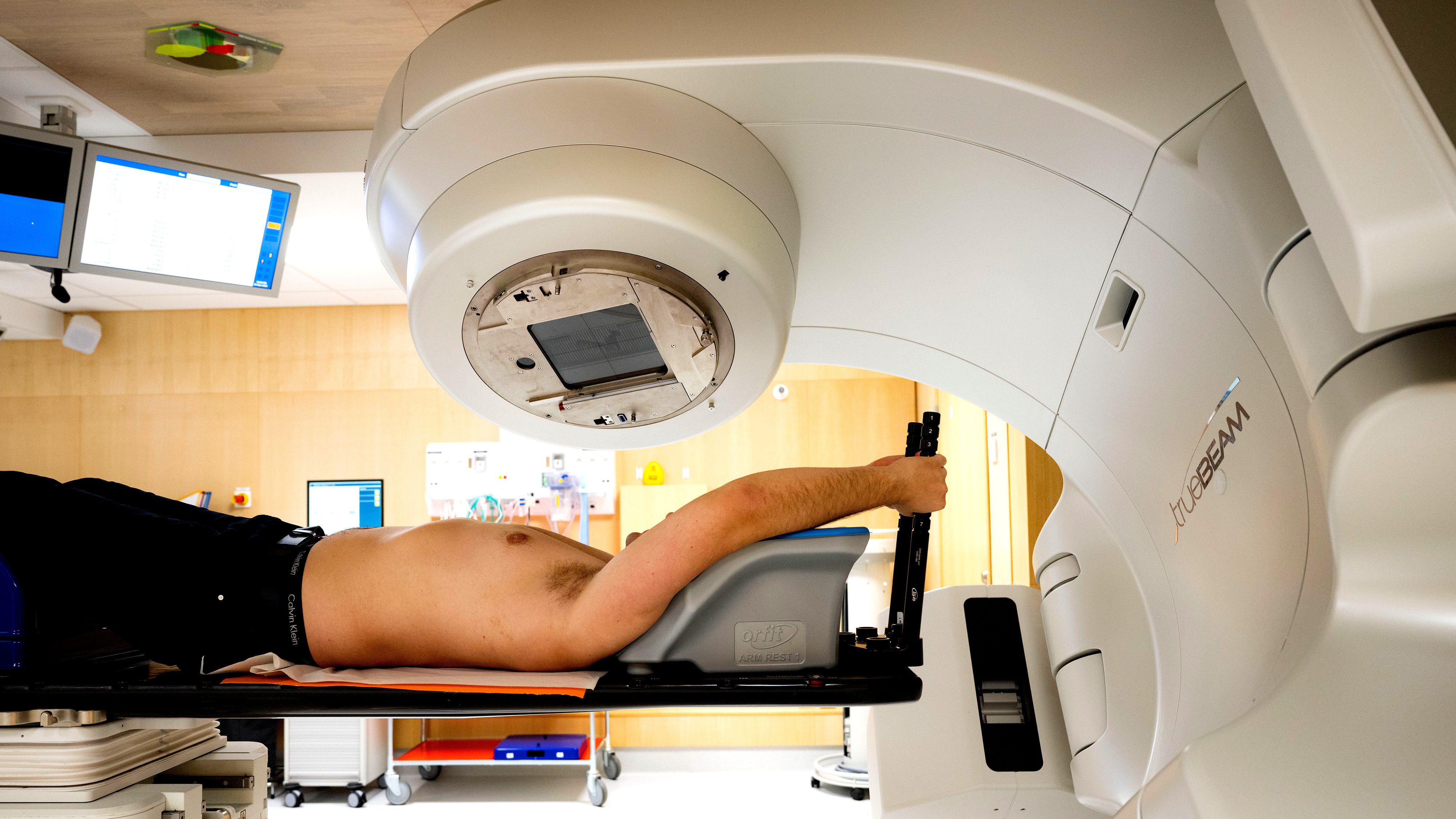 En person ligger på en brits under en stor medicinsk maskin i en vårdmiljö inför eller under strålbehandling.